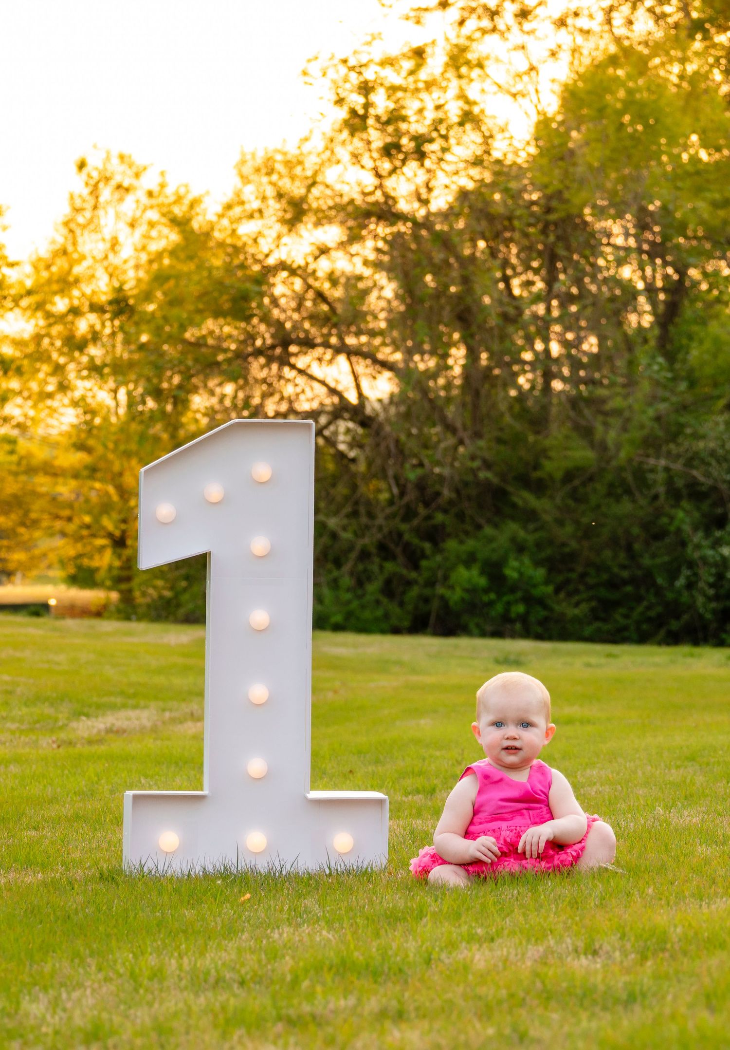 Baby girl in pink romper sits next to a large lit marquee number 1 on green grass.