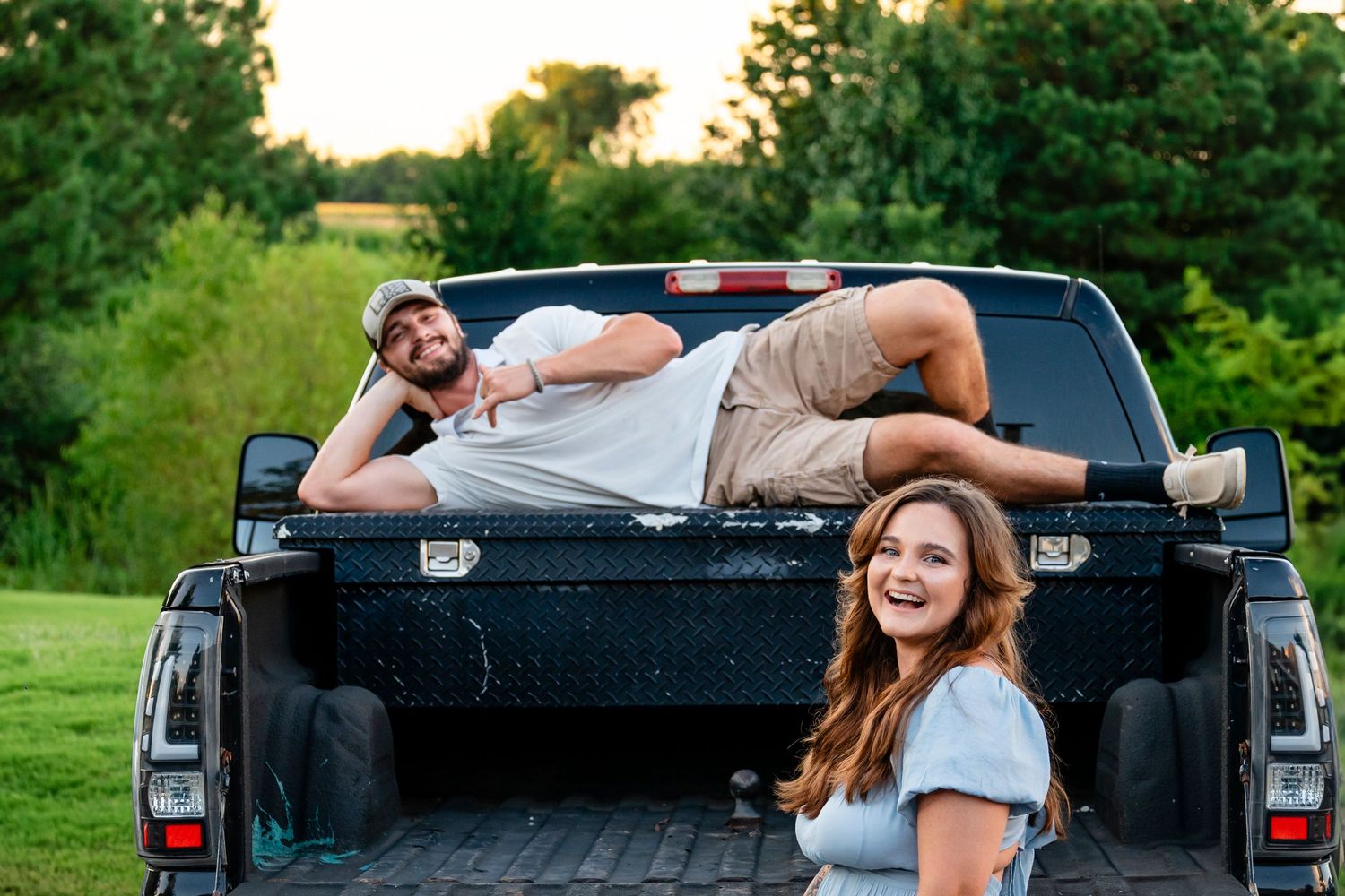A laughing woman stands by a truck as a man reclines in the bed at golden hour outdoors.