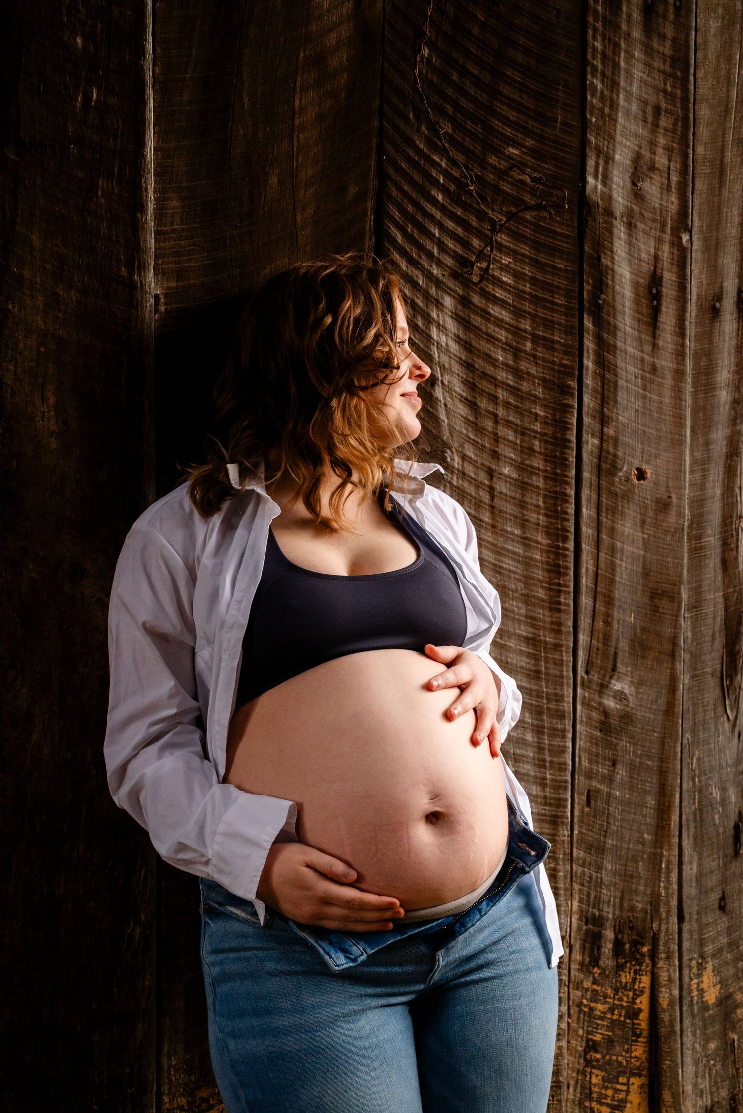 Pregnant woman wearing glasses and open white shirt cradles bare belly in jeans against rustic wooden wall, maternity photo.