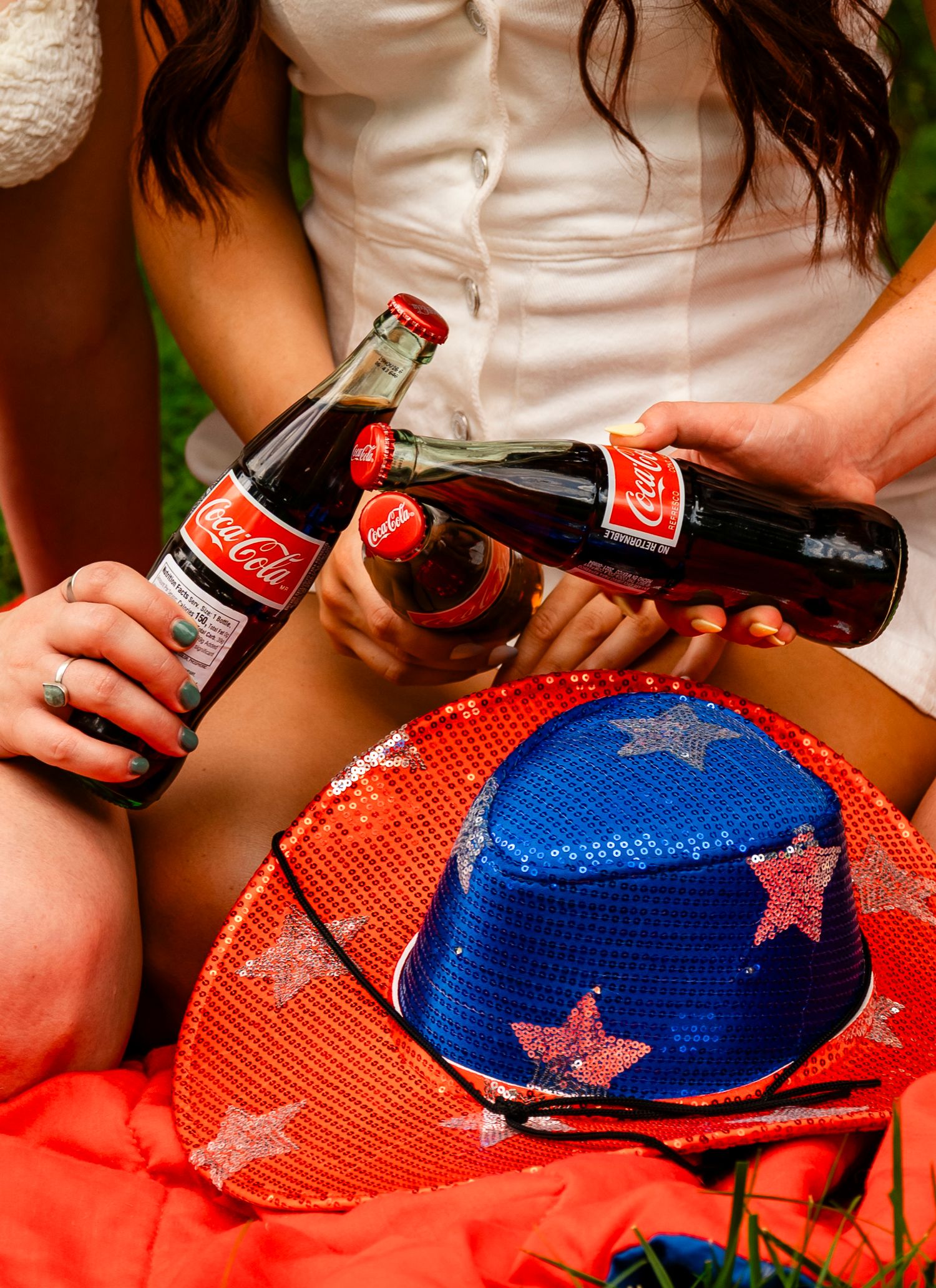 Two Coca-Cola glass bottles being clinked together near a red and blue sequin star-decorated hat outdoors.