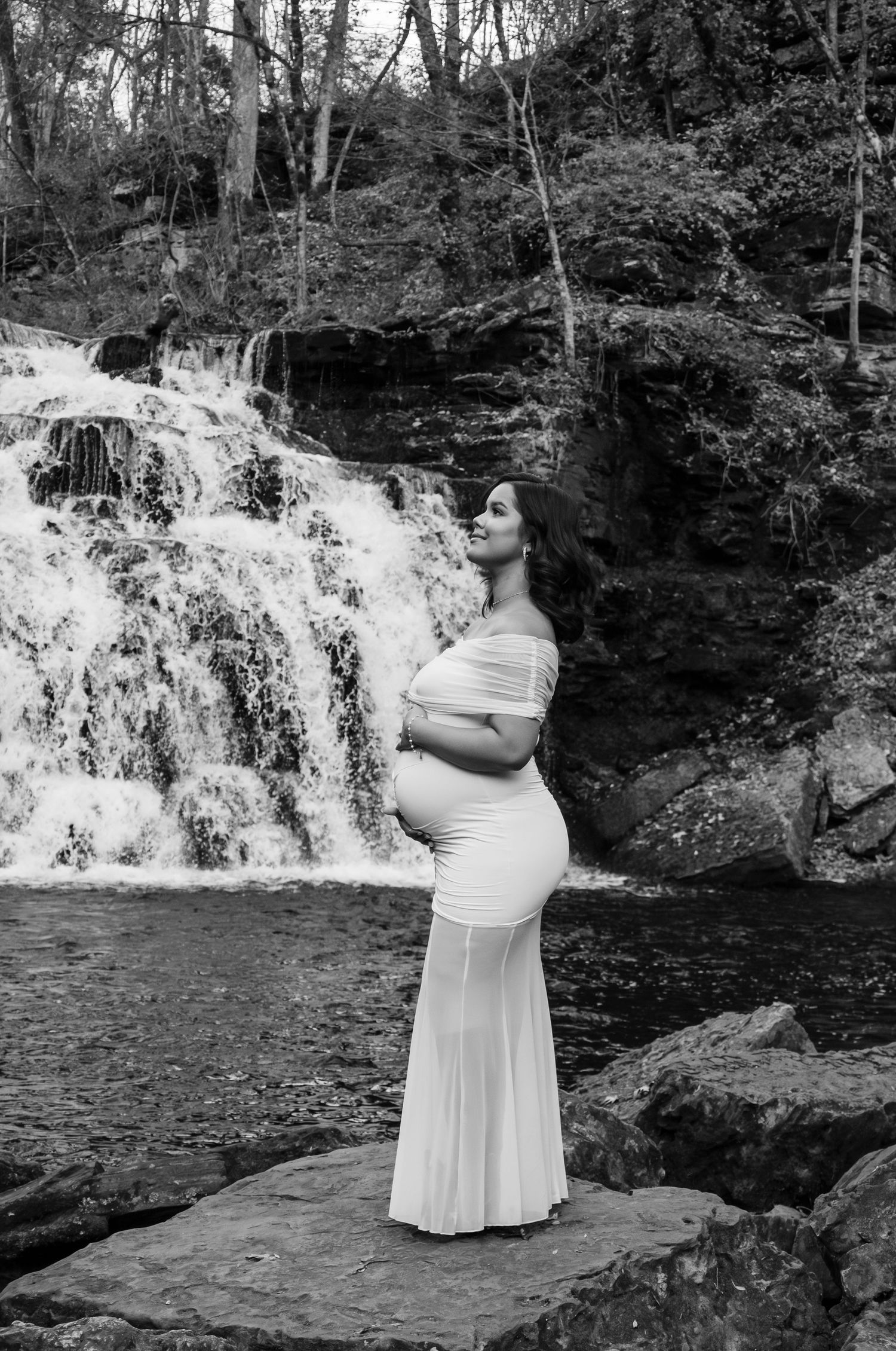 Pregnant woman in white off-shoulder gown poses gracefully before a cascading waterfall on rocky terrain.