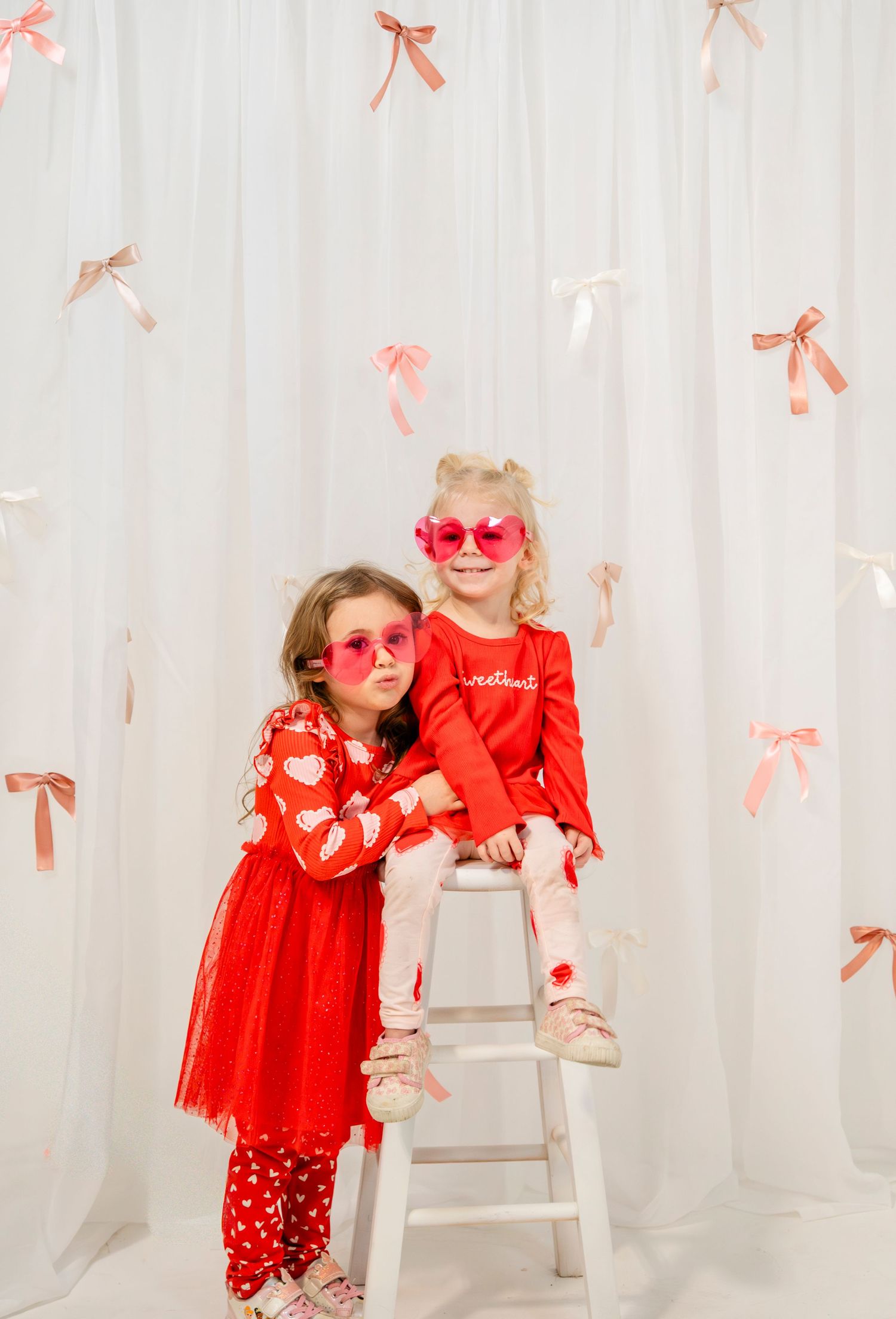 Two young girls in matching red Valentine's Day outfits wearing pink heart-shaped sunglasses pose together.