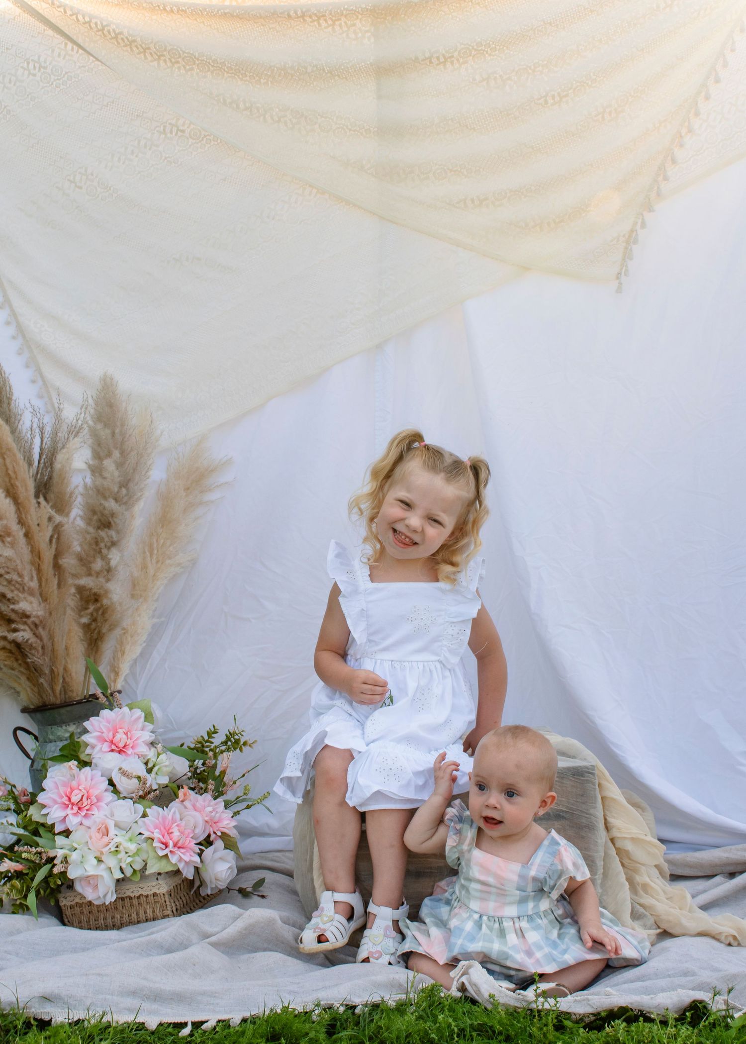 Two young sisters pose for a portrait, older girl in white dress with baby sibling among pink flowers and pampas grass.