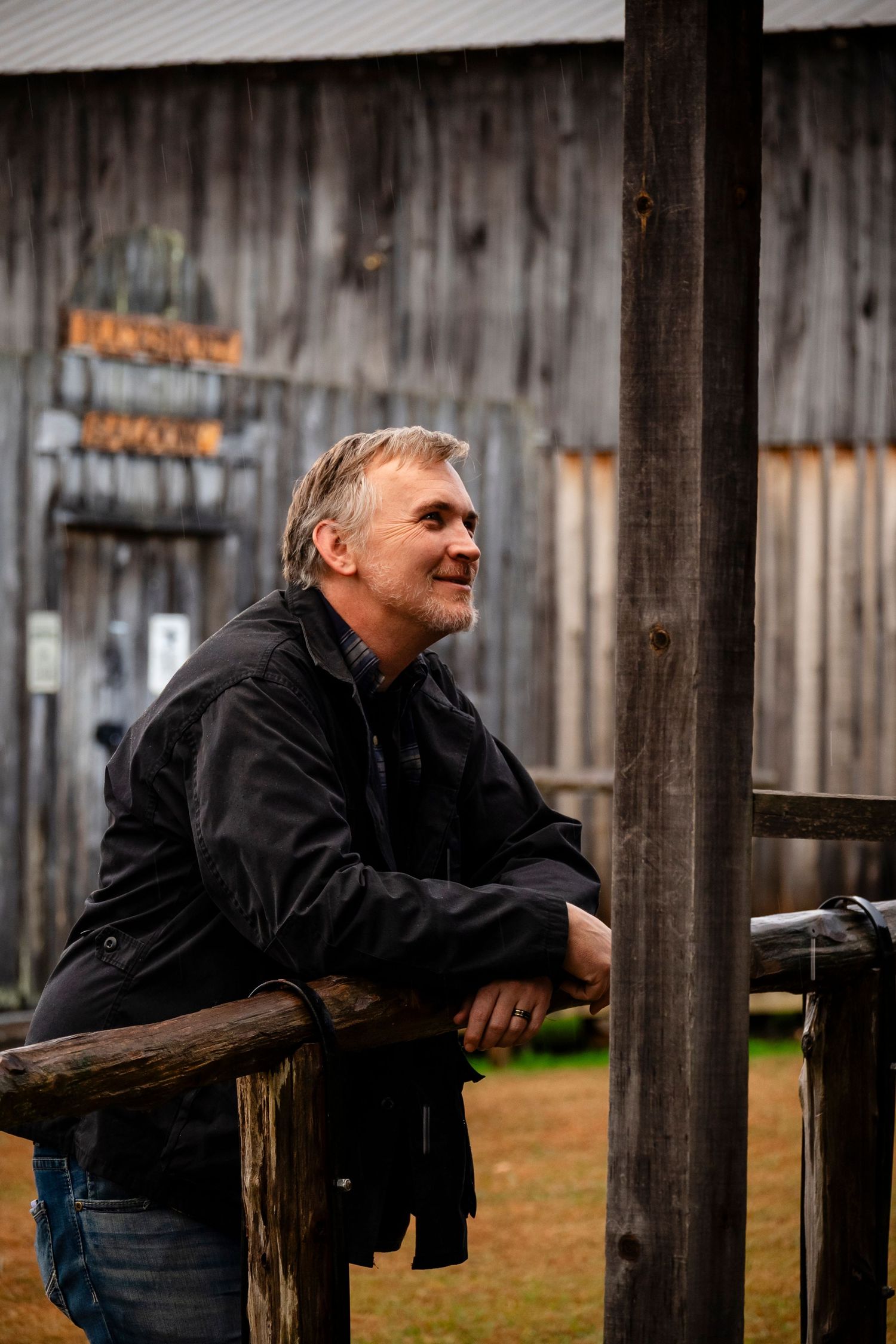 Middle-aged man in black jacket leaning on wooden fence, smiling outdoors near a weathered barn.