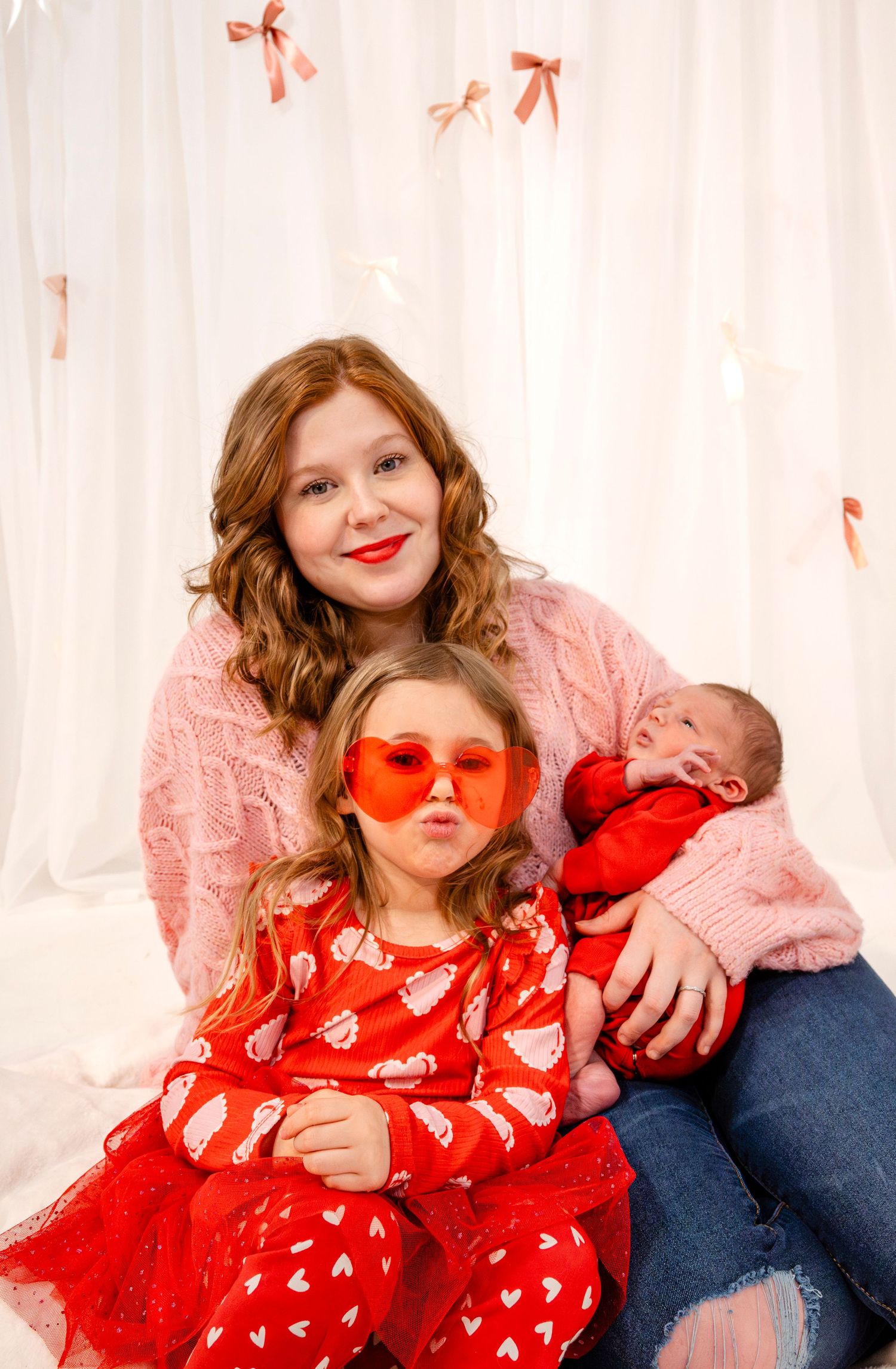 Redheaded mother in pink sweater poses with young daughter in red dress and newborn baby.