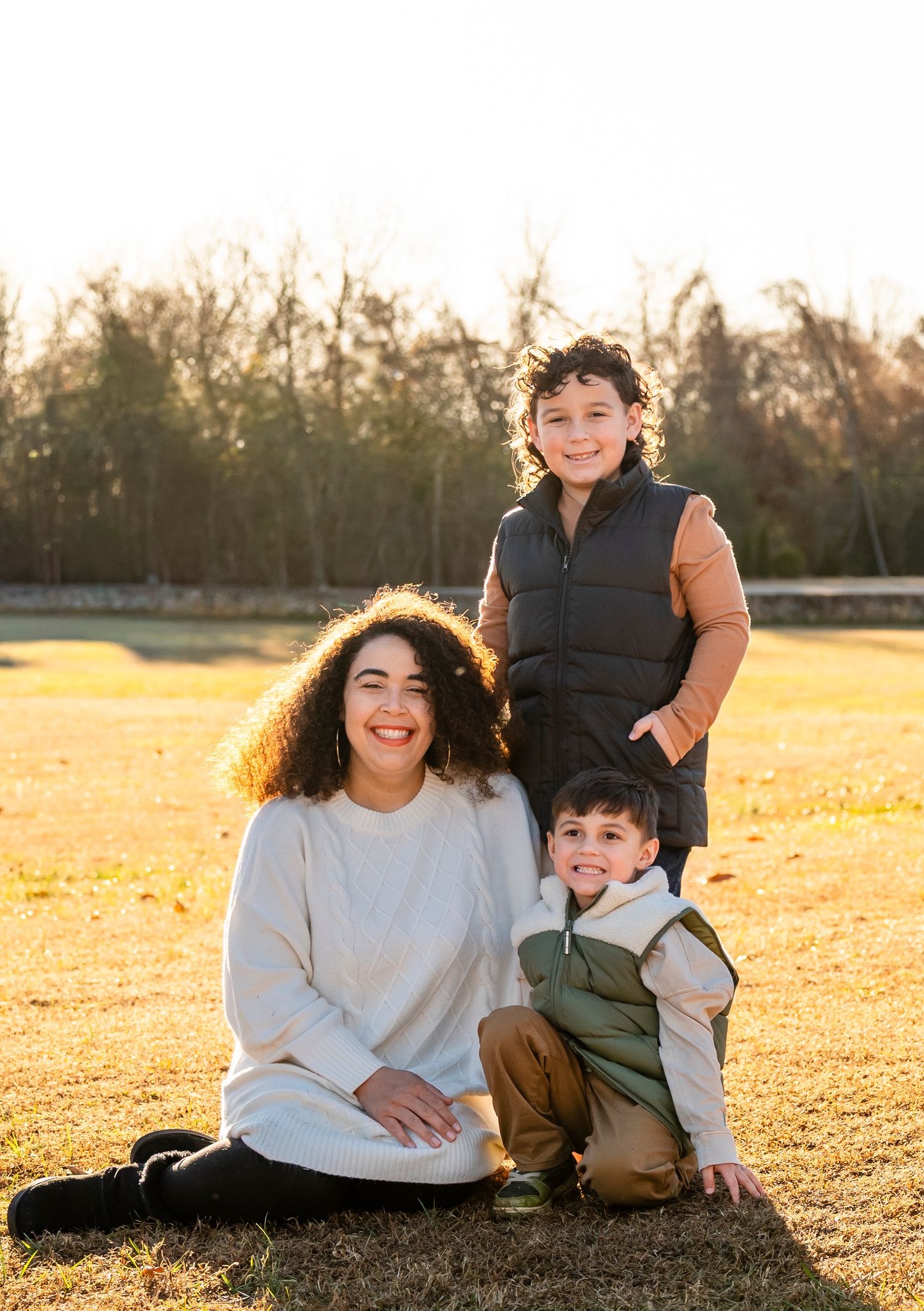 A mother with curly hair poses with her two smiling boys on a sunlit grassy field at golden hour.