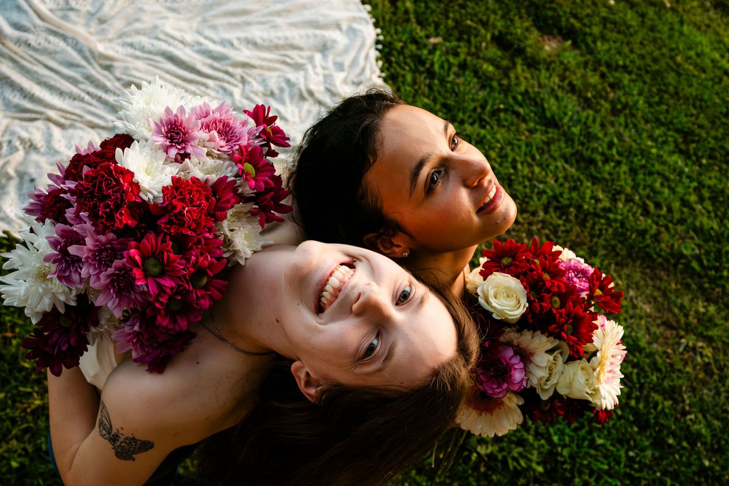 Two women lying on grass with colorful floral bouquets of red, pink, and white flowers, smiling upward.