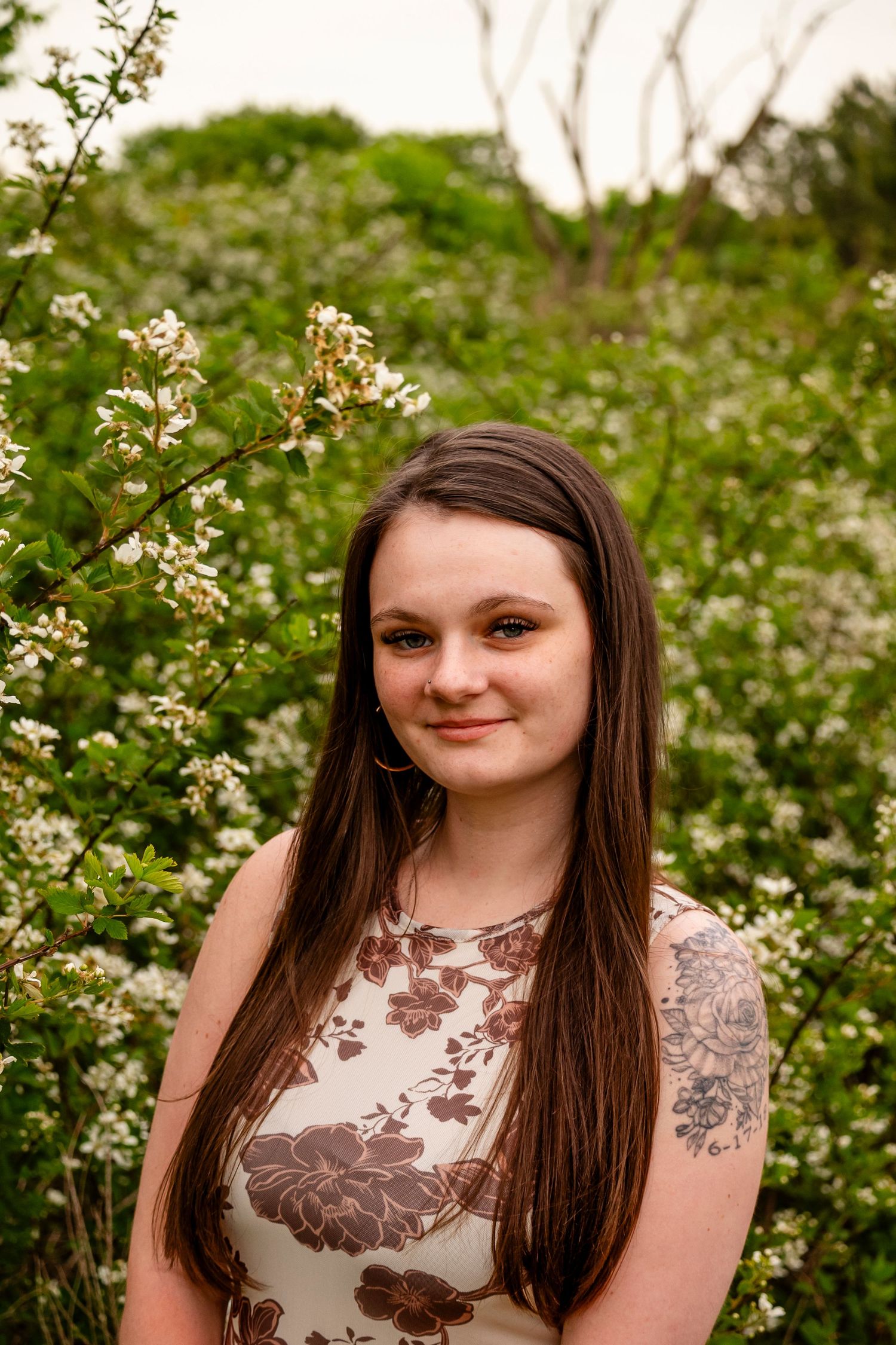 Young woman with long brown hair and floral tattoo smiles in front of blooming white wildflowers outdoors.