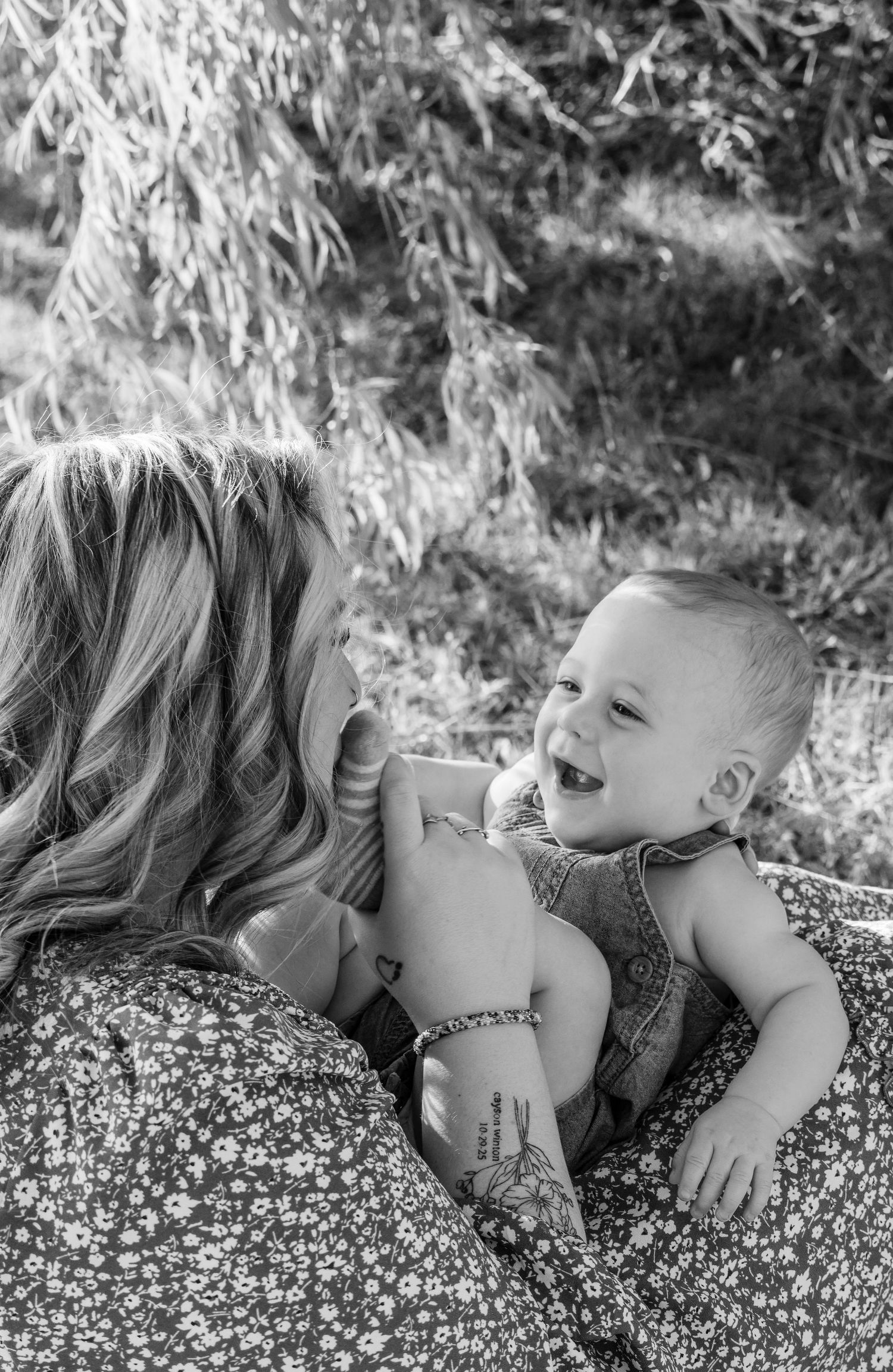 Black and white photo of a laughing baby being held by a blonde woman outdoors near a willow tree.