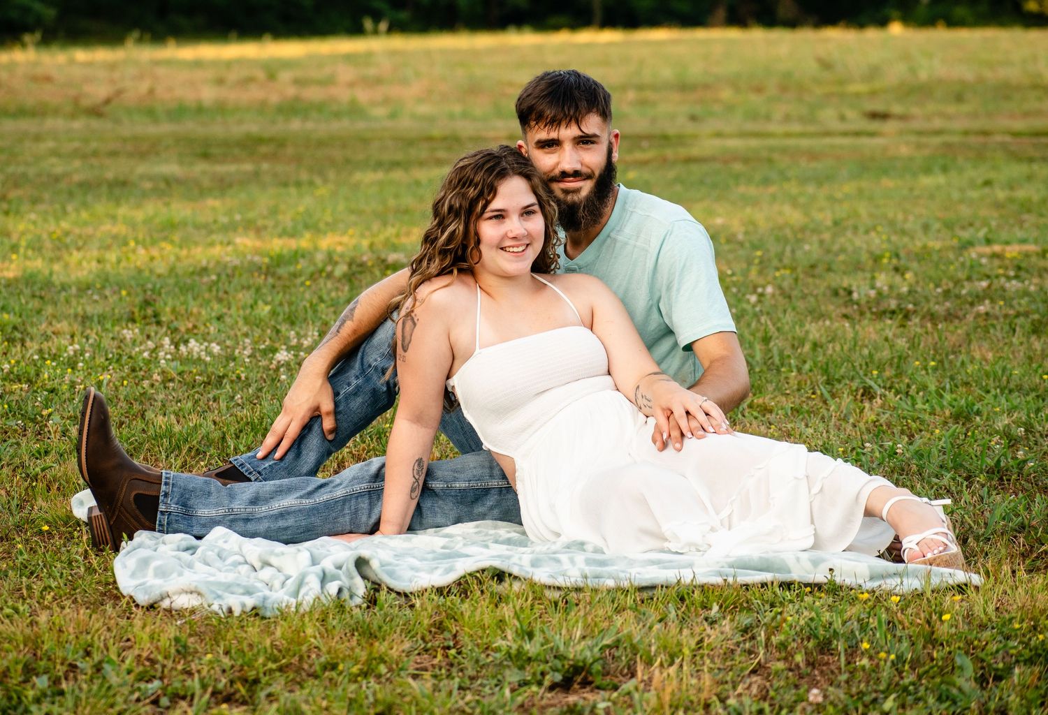 Expecting couple sitting on blanket in green field, woman in white maternity dress leaning against partner.