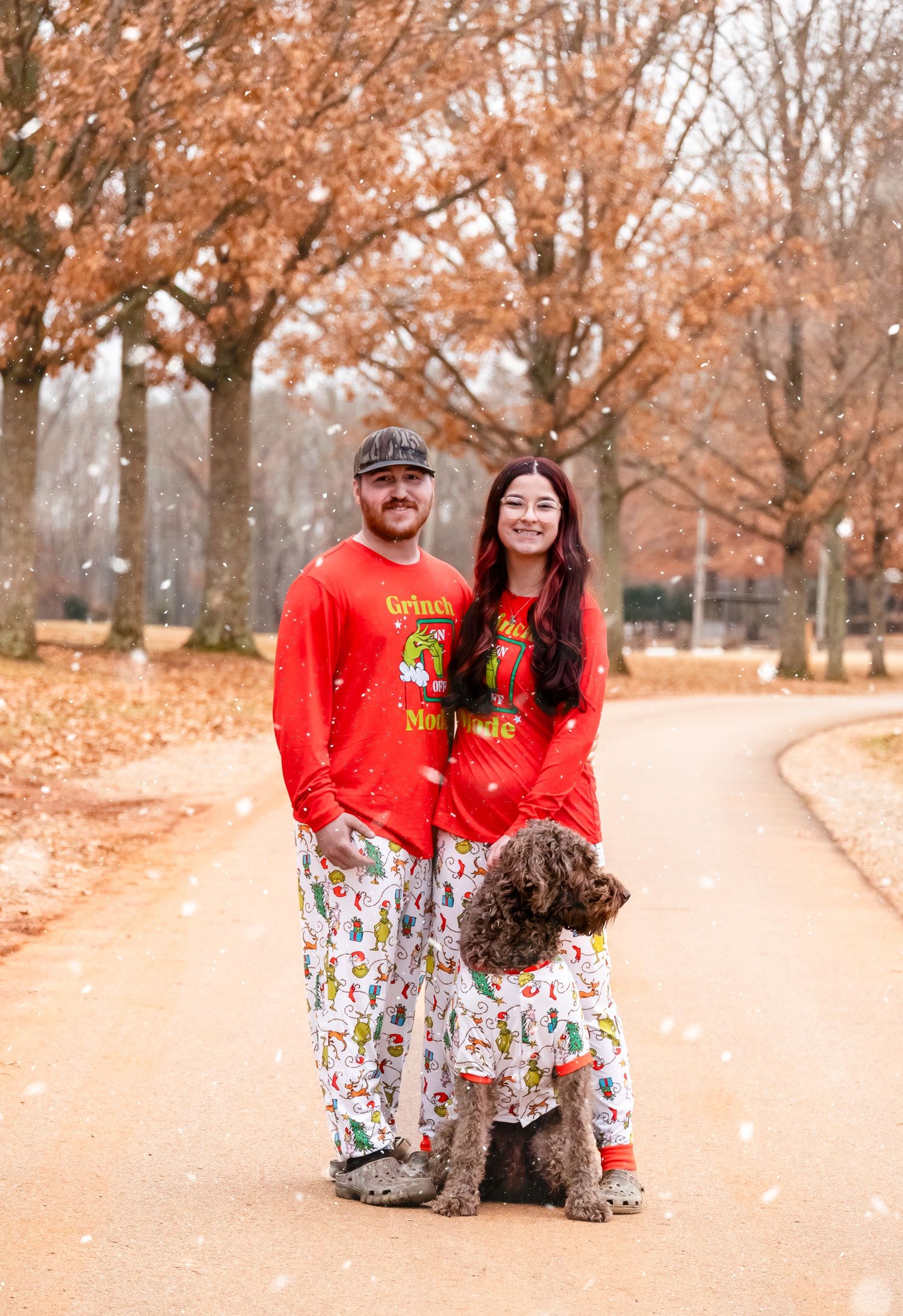 Couple in matching Grinch Christmas pajamas poses with their curly dog on a tree-lined path.