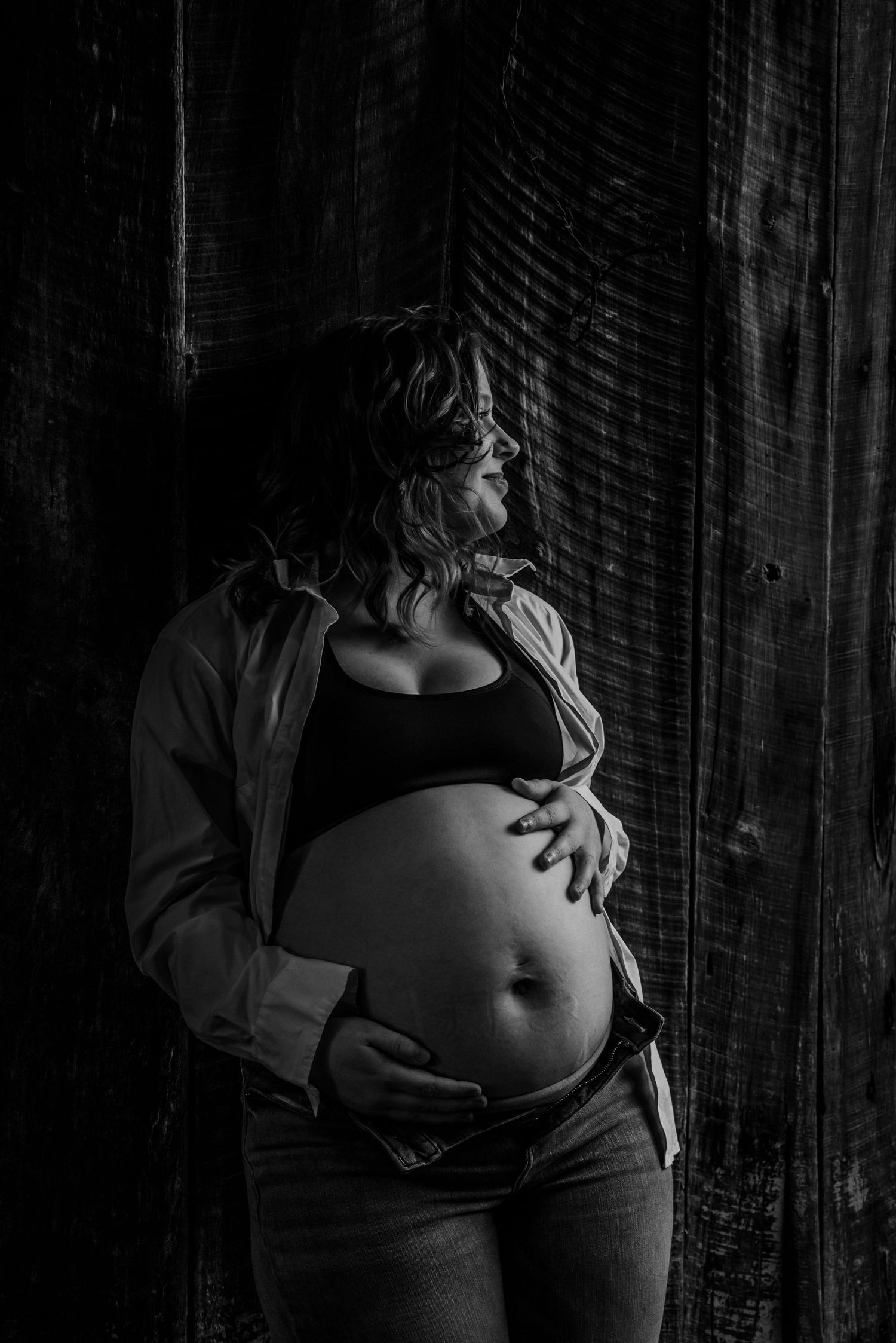 Dramatic black and white maternity photo of pregnant woman cradling baby bump against dark rustic wooden wall.