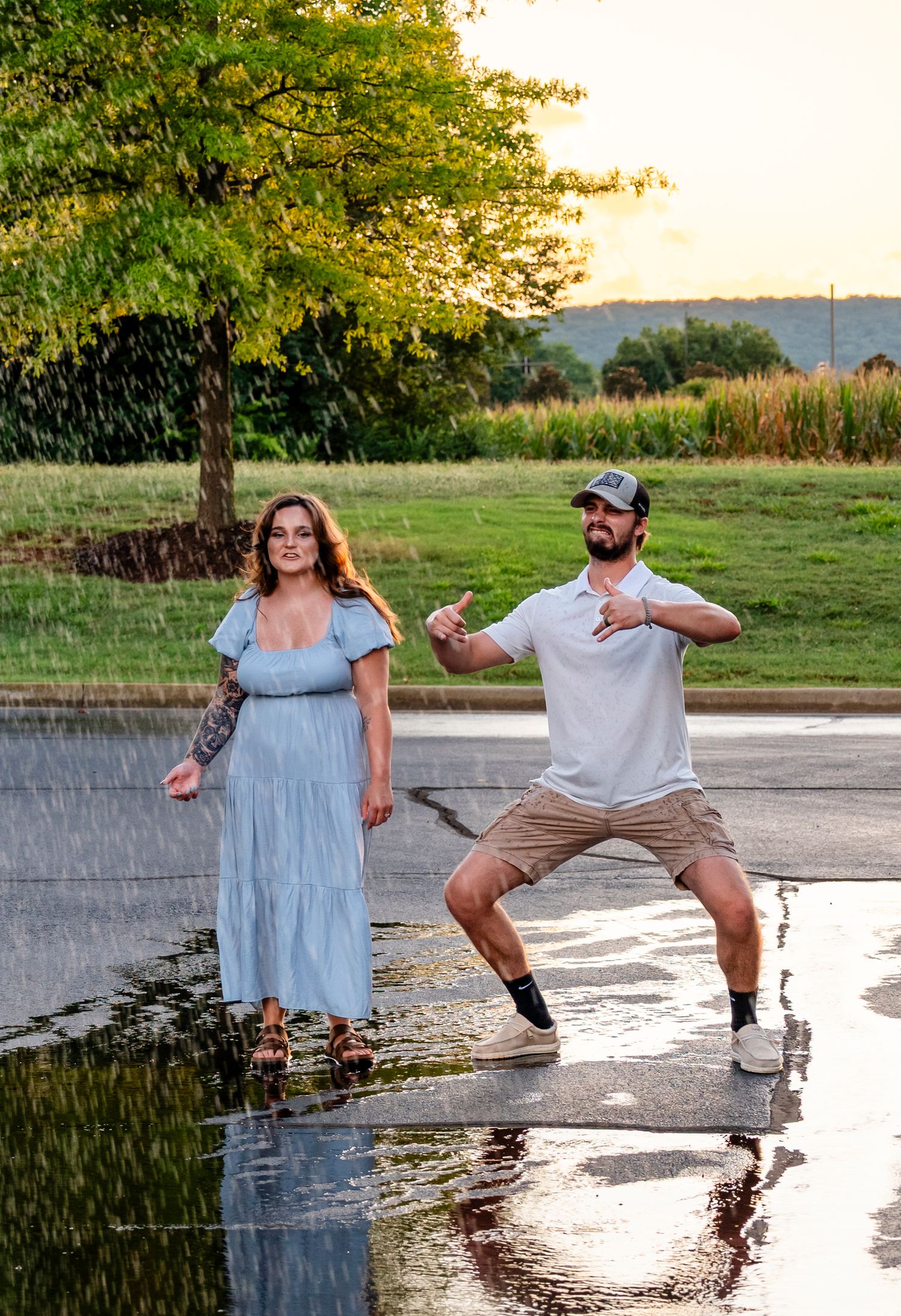 A couple poses playfully in the rain at sunset, woman in blue dress, man doing a fun stance.