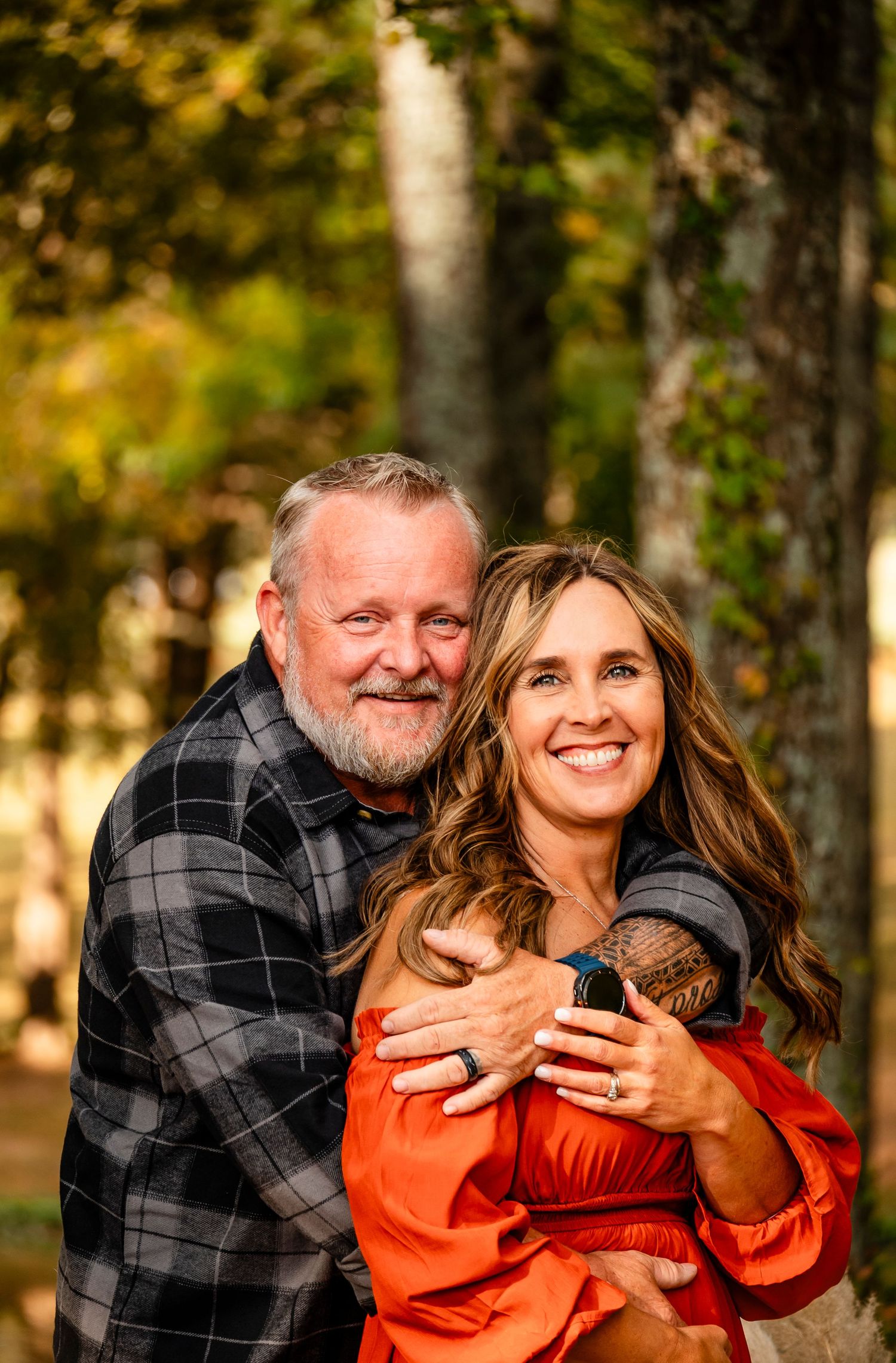 A smiling couple embraces outdoors in autumn, woman in orange dress holding a newborn baby, man in plaid shirt behind her.