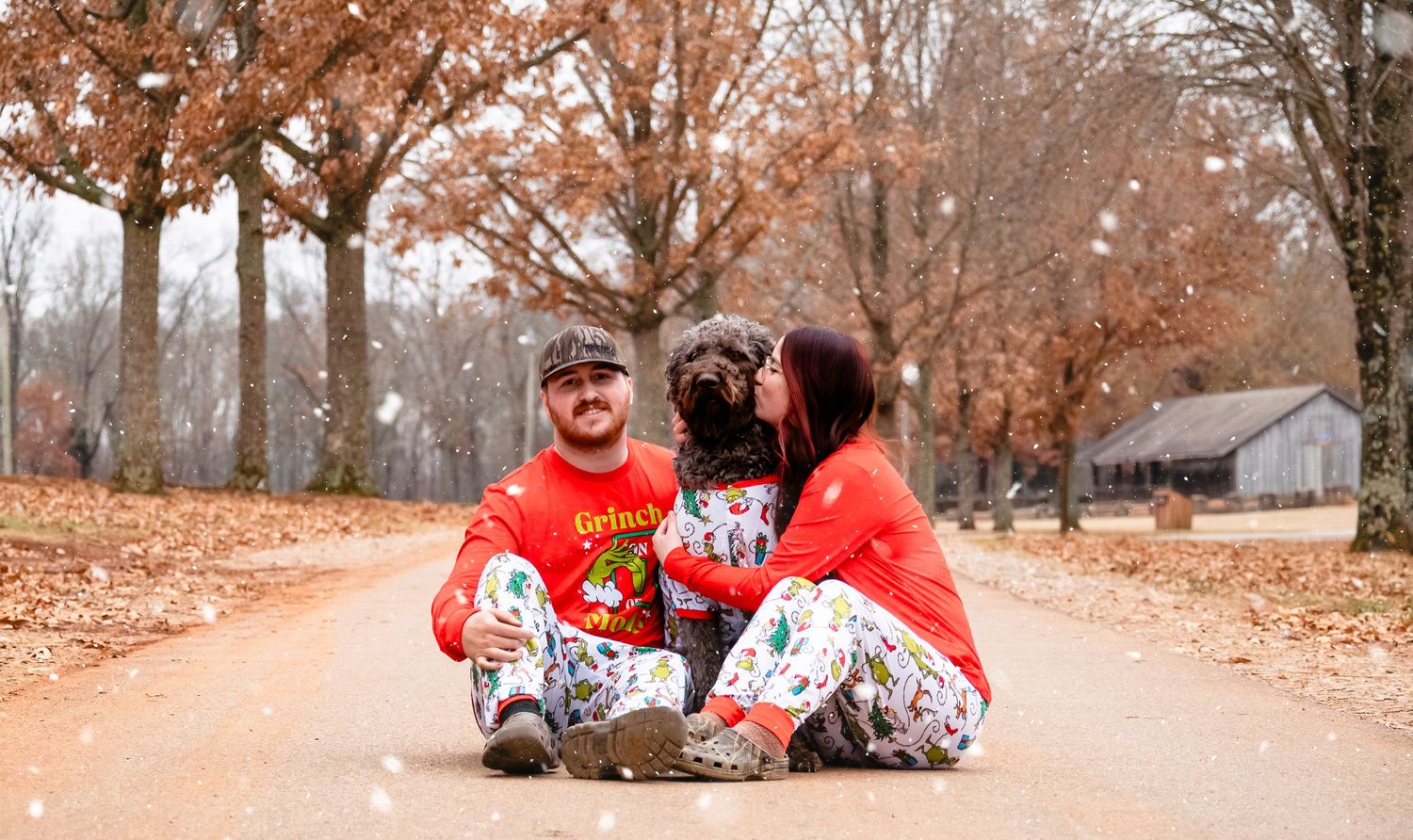 Couple in matching Christmas pajamas share a kiss with their dog on a scenic outdoor path.