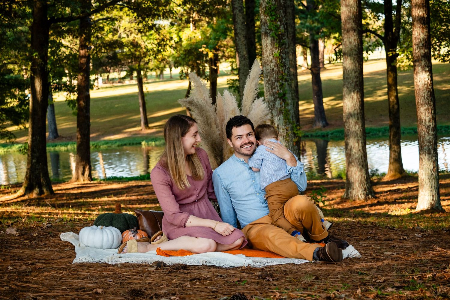 Young family of three relaxing on blanket in sunny autumn park by pond.