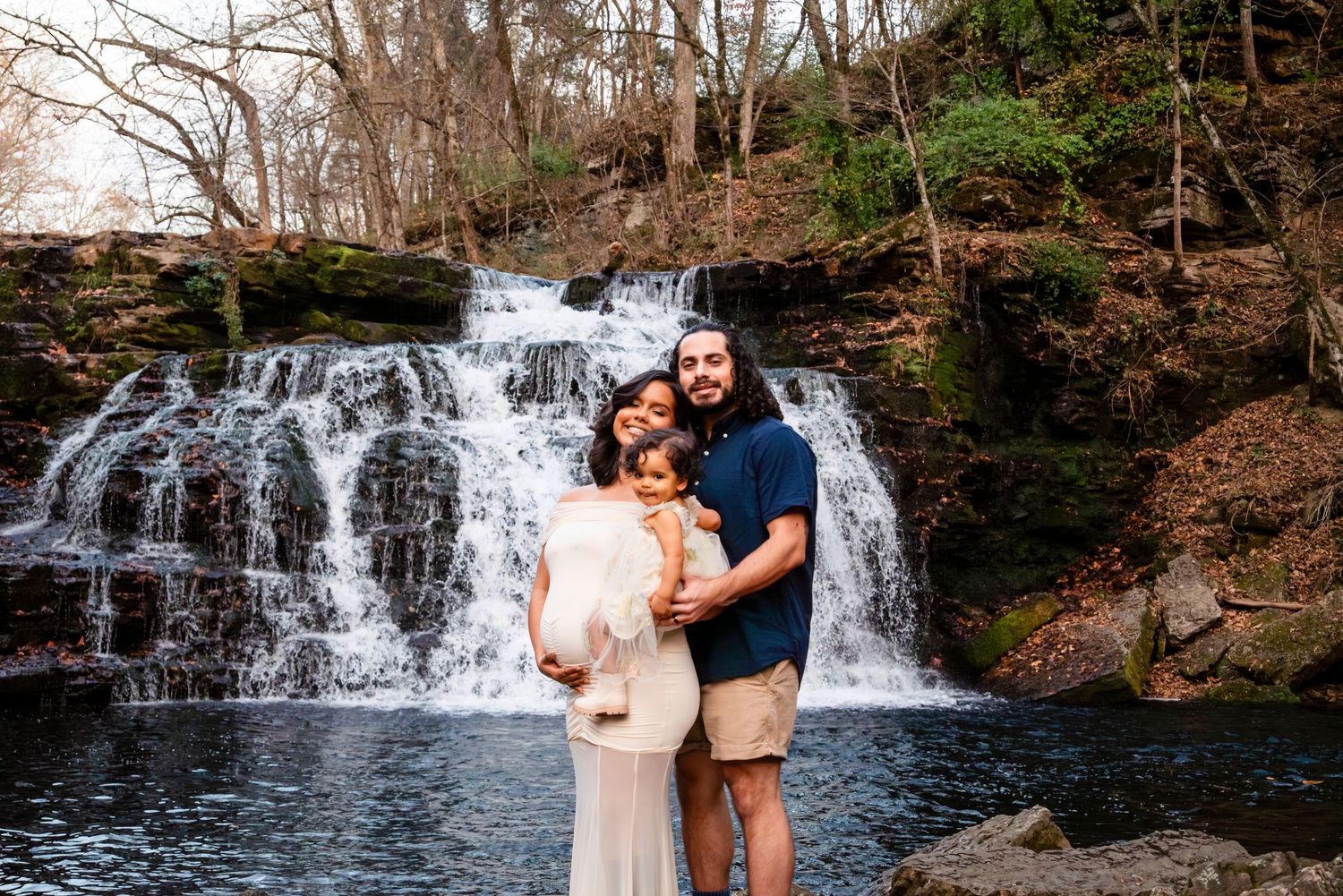 Couple holding toddler poses together in front of a scenic cascading waterfall in a wooded setting.