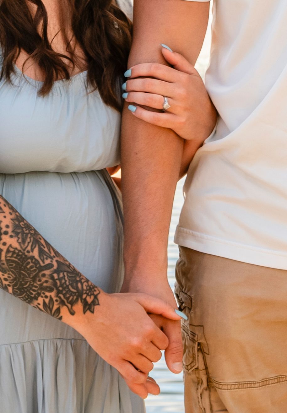 Close-up of a couple holding arms, woman with floral tattoo and engagement ring, soft outdoor light.