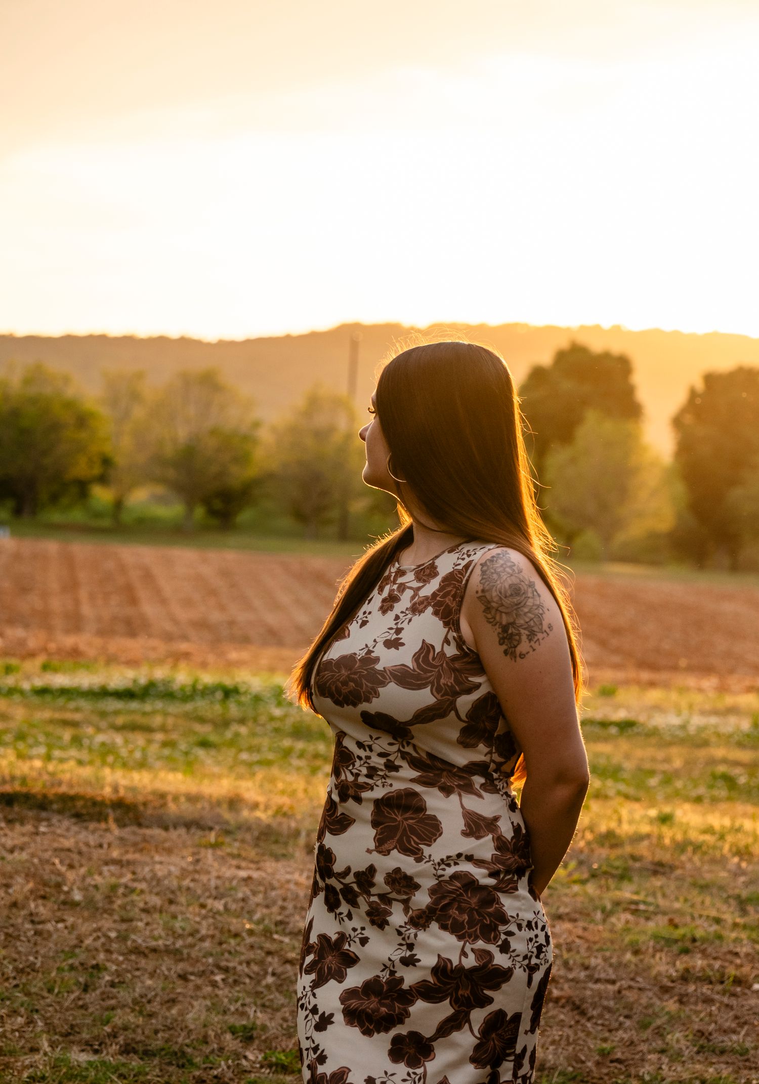 Woman in floral dress standing in golden sunset field, gazing at scenic rural landscape with trees and hills.
