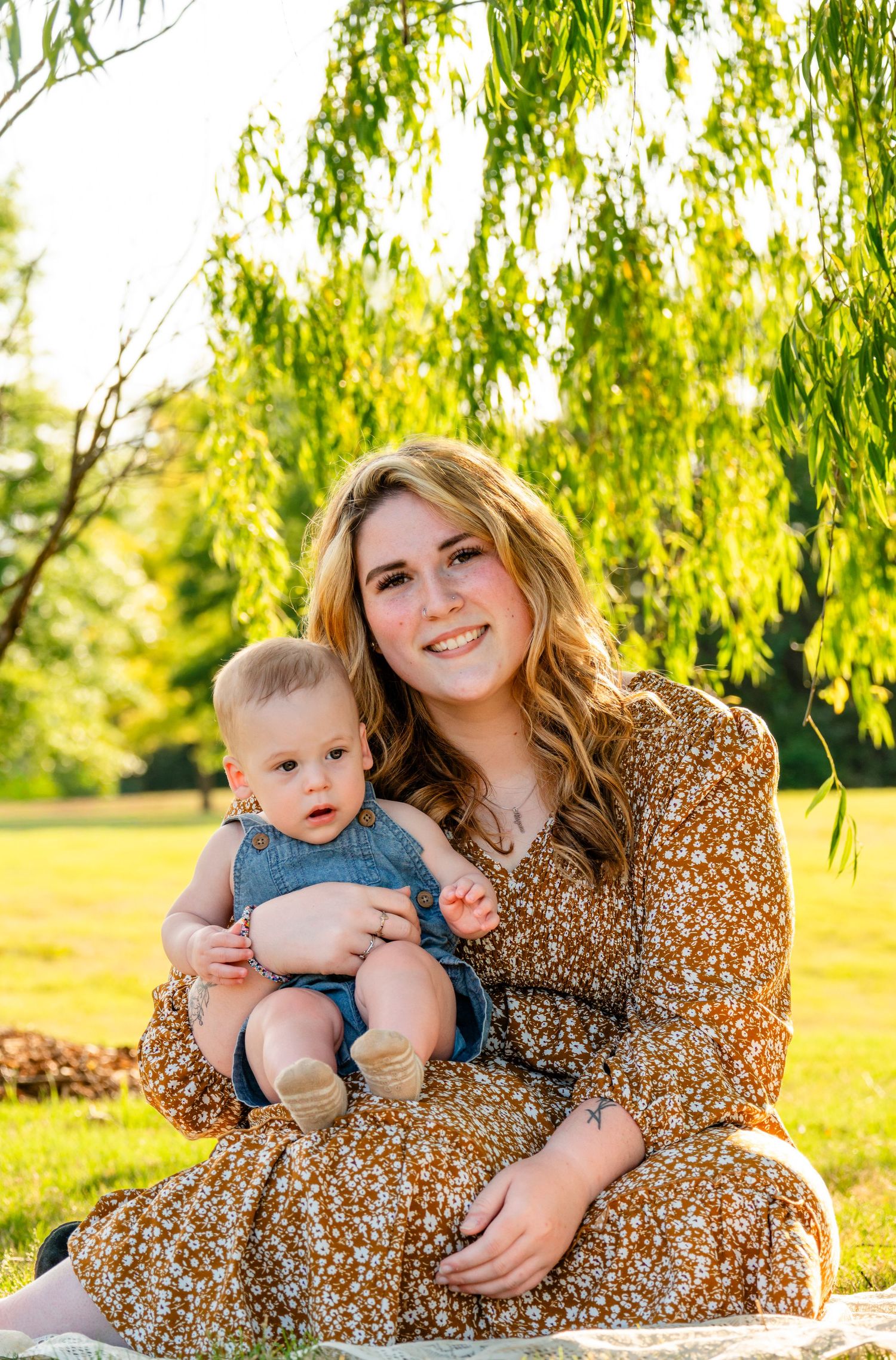 Young woman in floral dress holds baby boy outdoors near willow tree on sunny day.