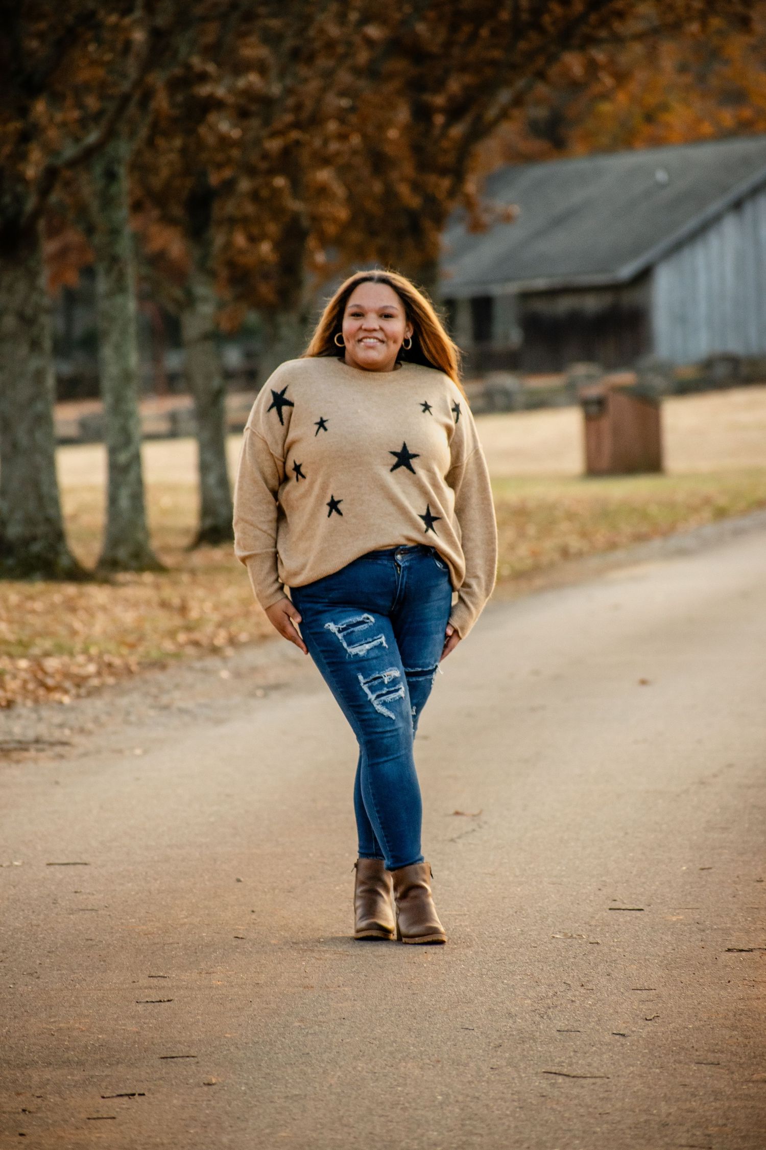 Woman in star-print sweater and ripped jeans walks down a rural path surrounded by autumn trees.