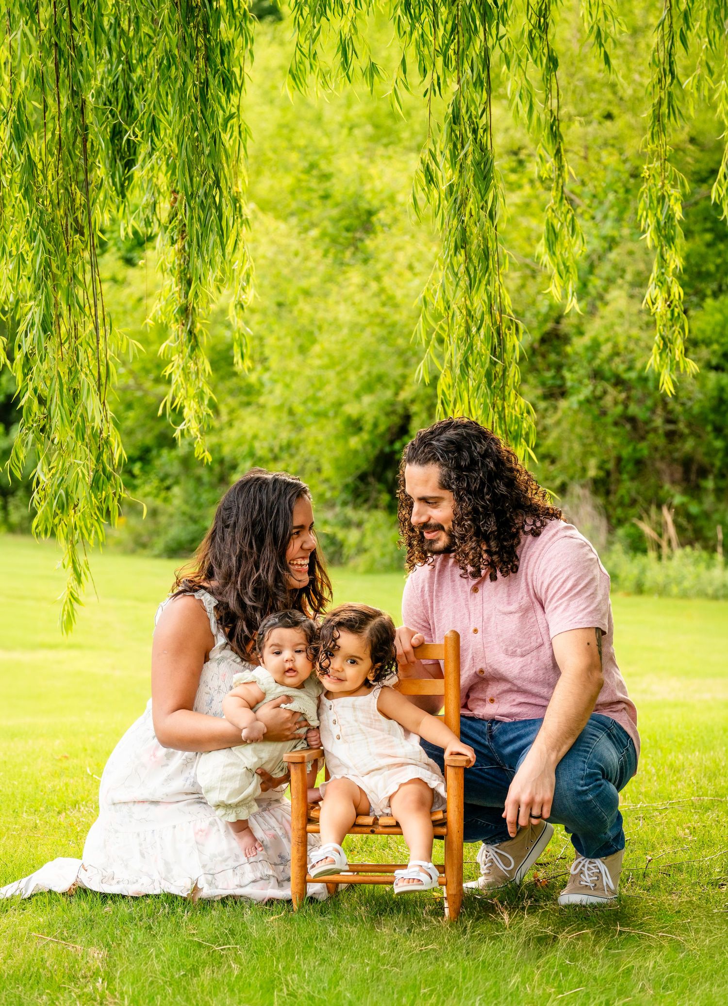 Family of four poses under a weeping willow tree on green grass, parents smiling at two young toddlers on a small wooden chair.