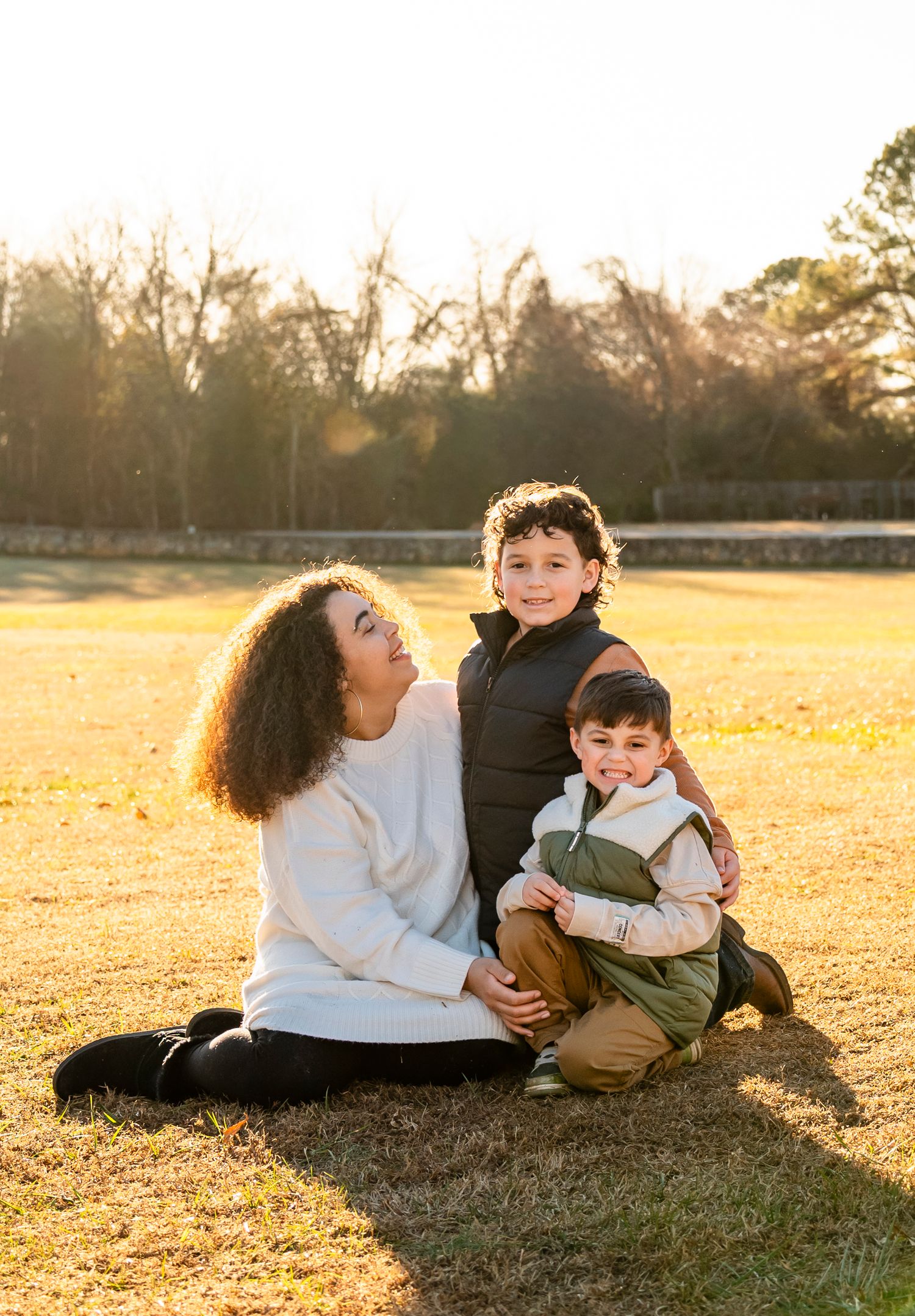 A mother sits on golden grass at sunset with her two young sons in a sunlit park setting.
