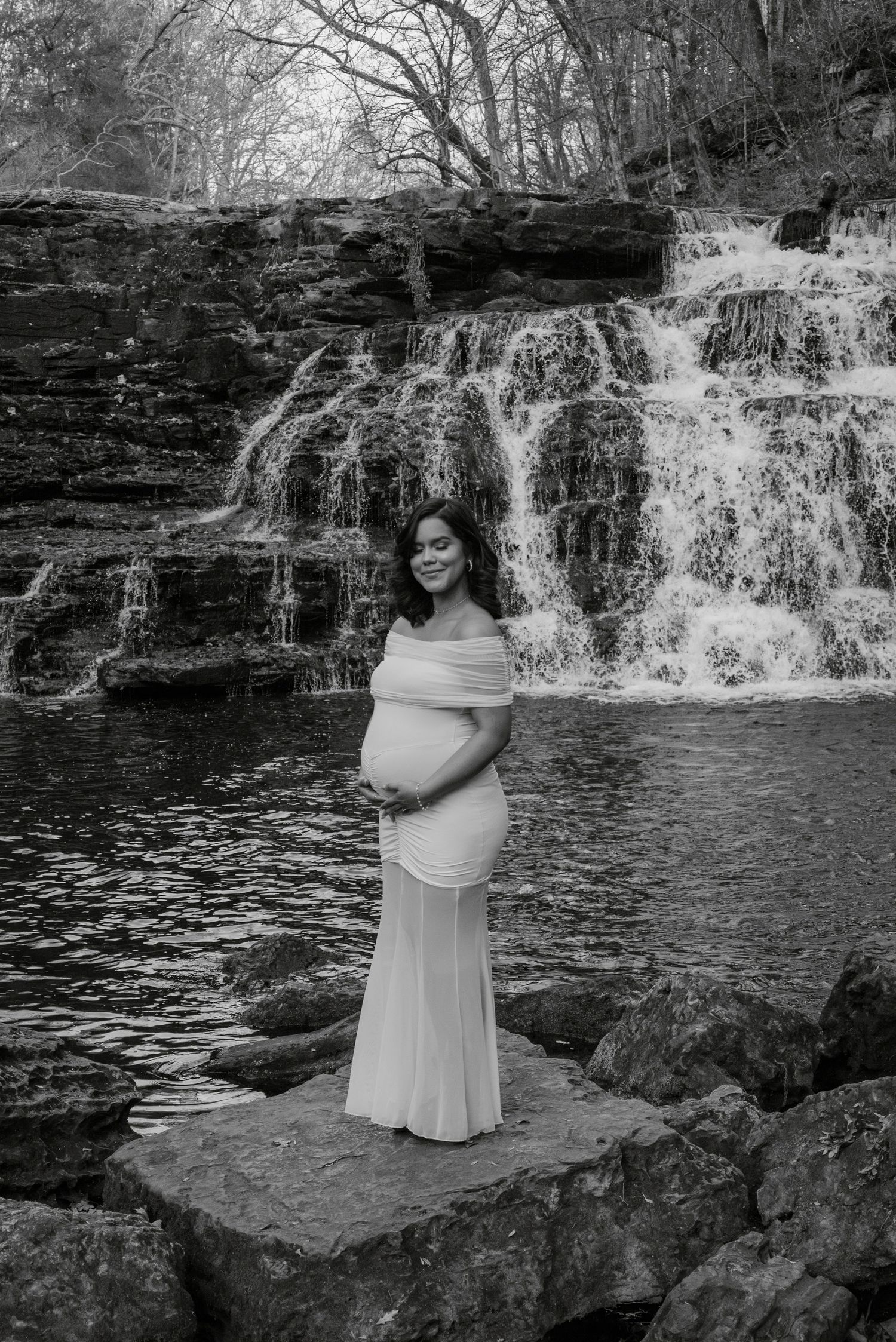 Pregnant woman in white off-shoulder gown poses gracefully before a cascading waterfall in black and white.