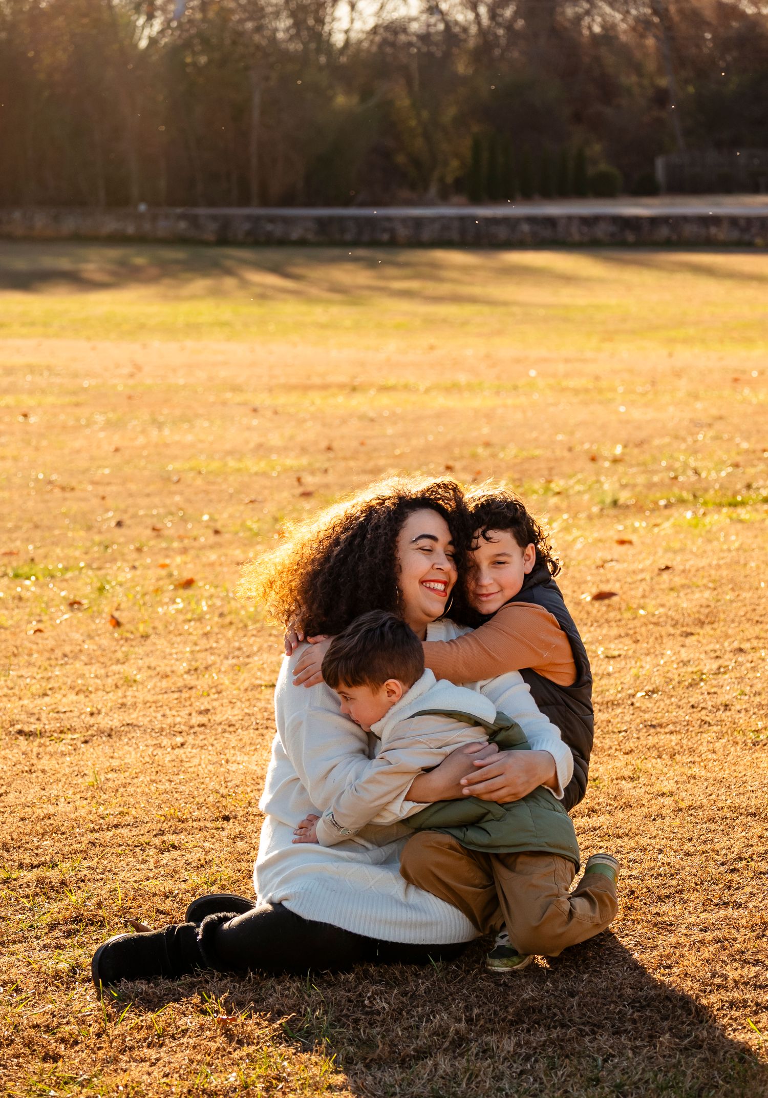 Mother sits on golden autumn grass hugging her two young sons in a warm sunset family portrait.