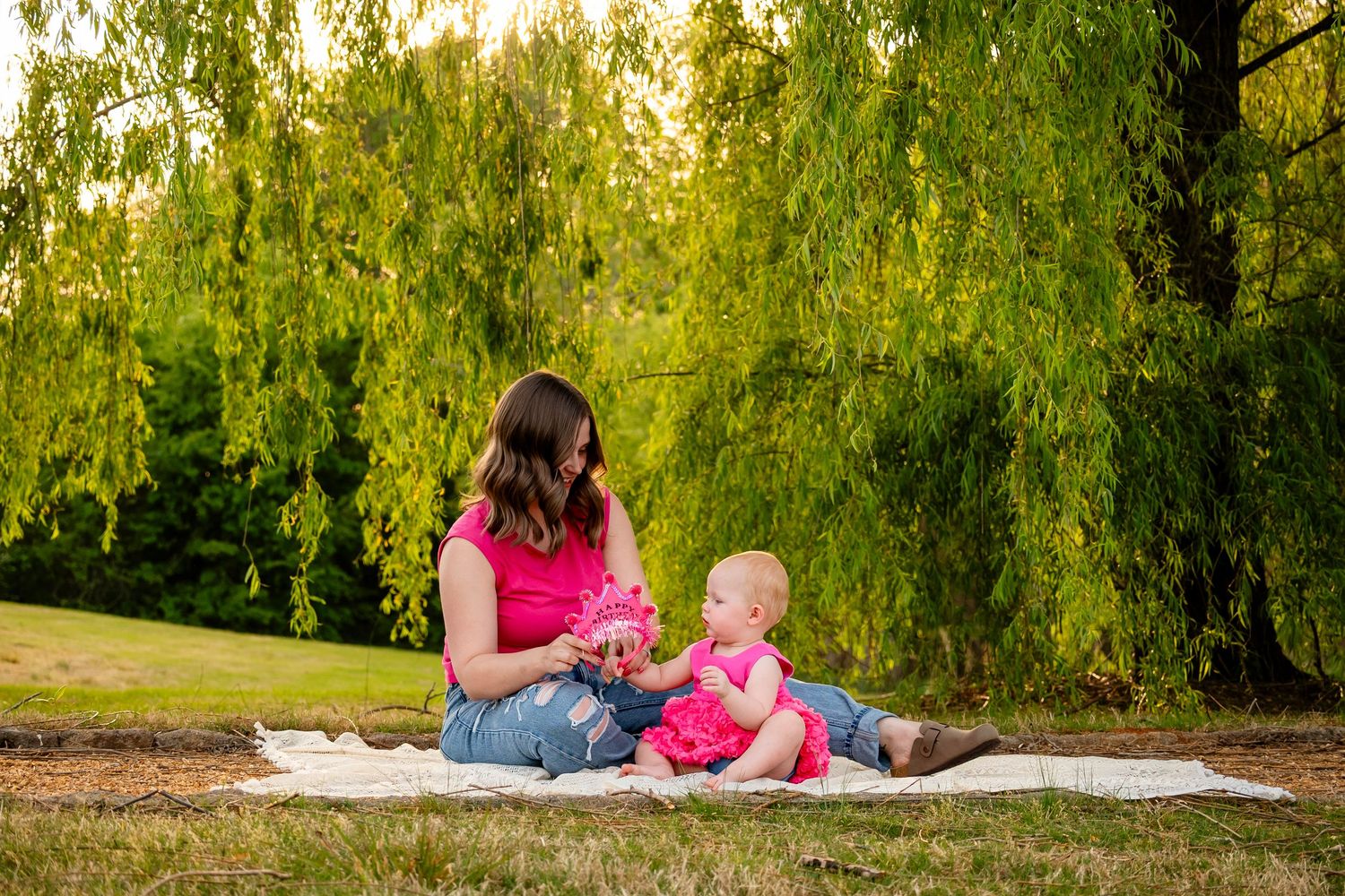 Mother in pink top sits on blanket with baby girl in pink dress under a weeping willow.