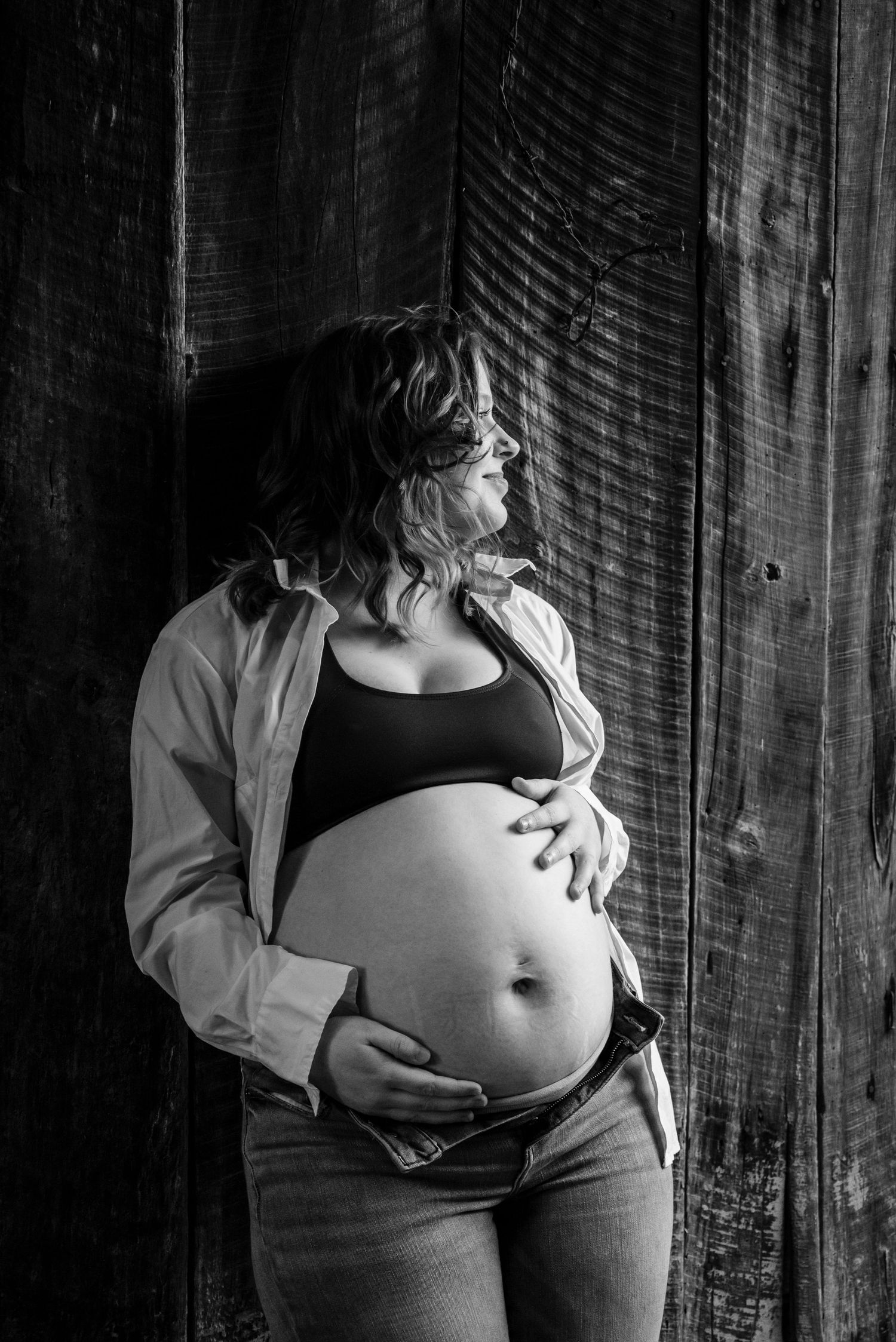 Black and white maternity portrait of pregnant woman in open shirt and jeans leaning against wooden barn wall.