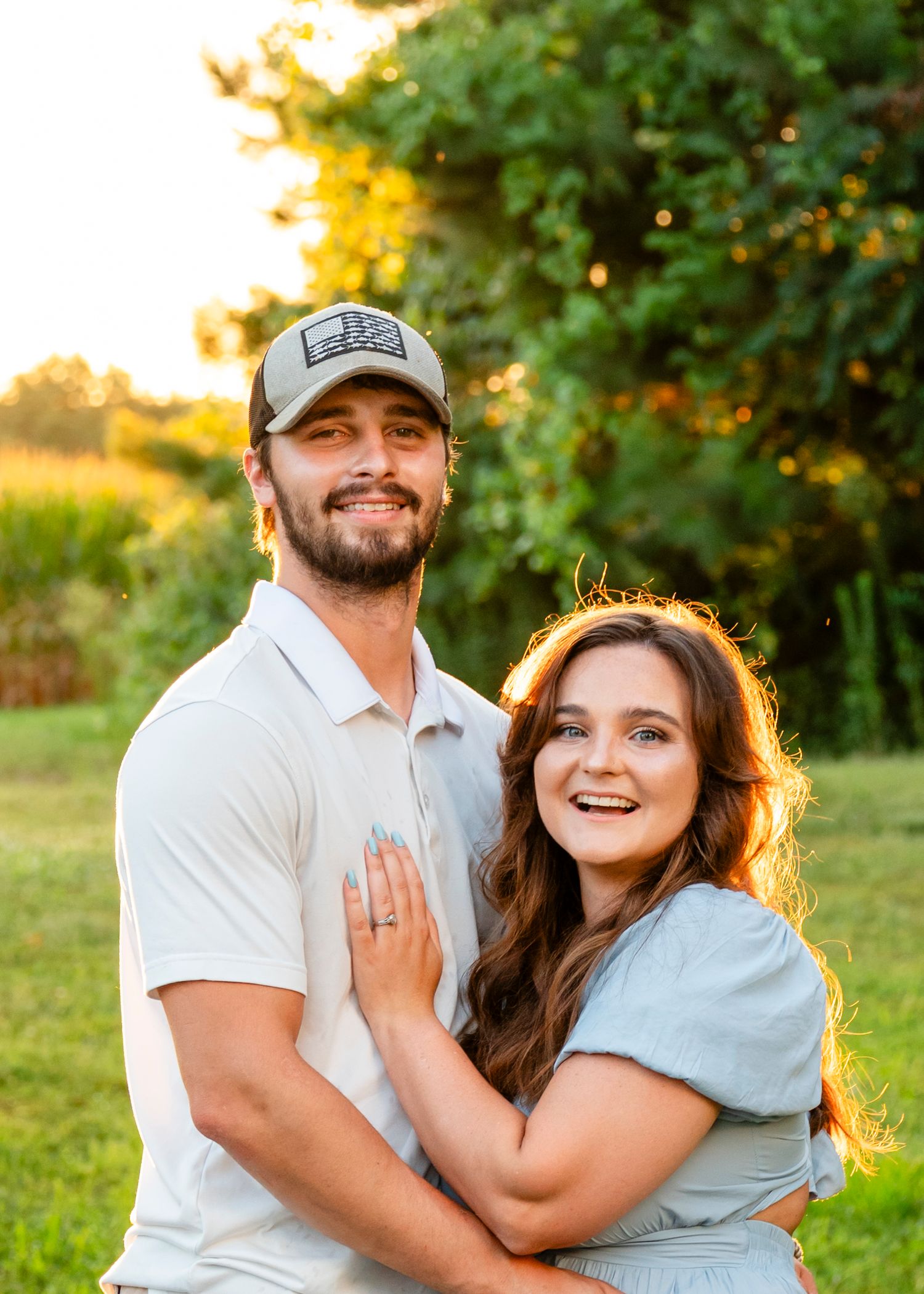 A happy couple embraces in a sunlit field, woman showing an engagement ring, both smiling brightly.