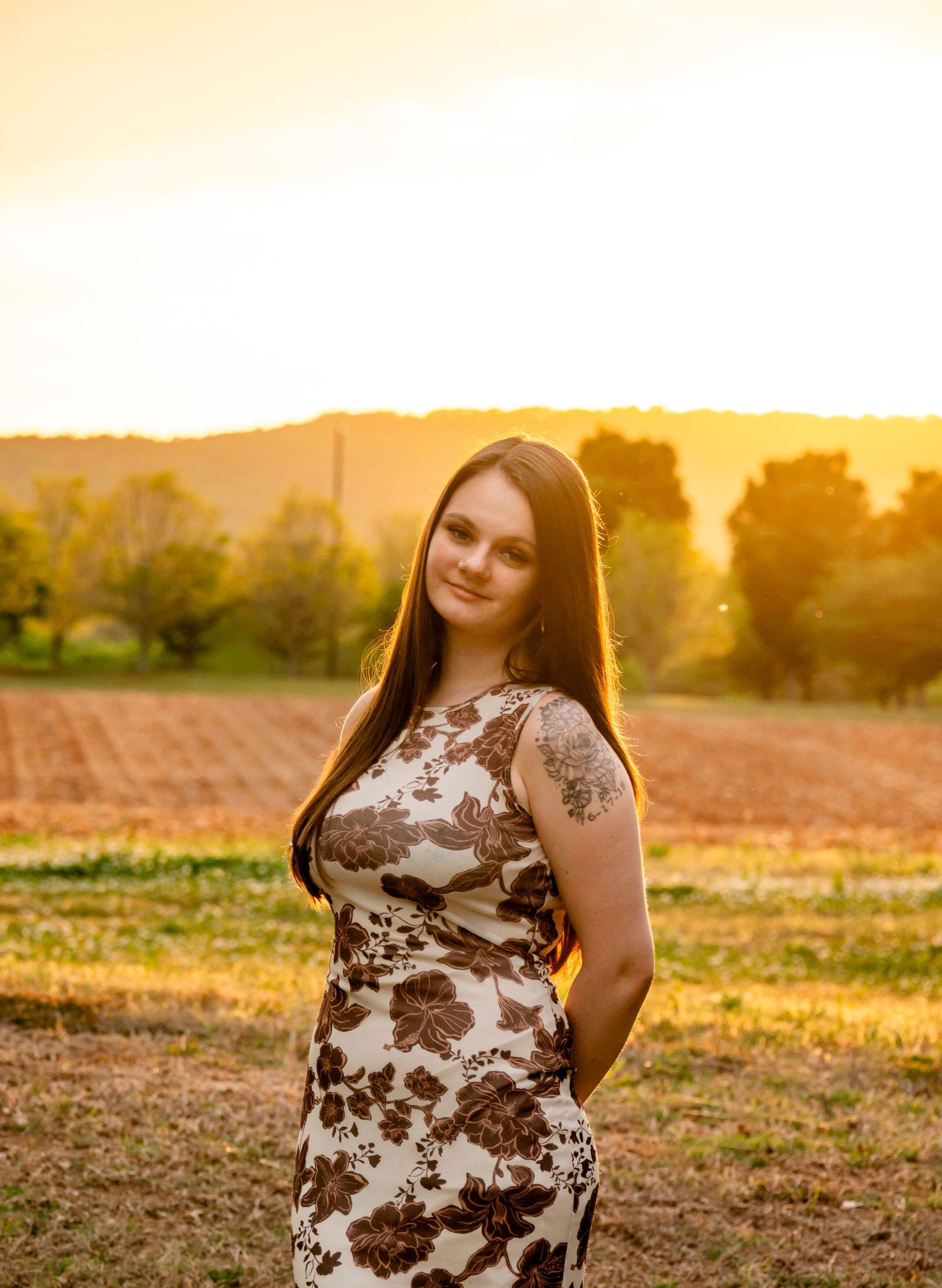 Young woman in floral dress poses in golden sunset light with rural farmland and trees in background.