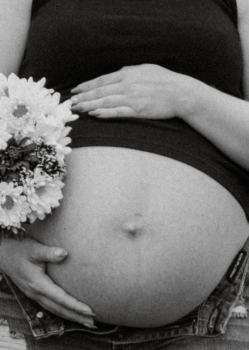 Black and white close-up of pregnant belly with white daisy flowers held gently by mother's hands.