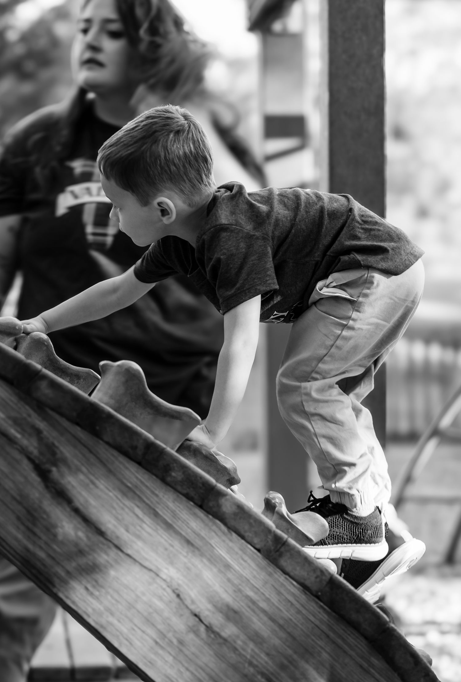 Black and white photo of young boy climbing wooden playground slide with woman in background.