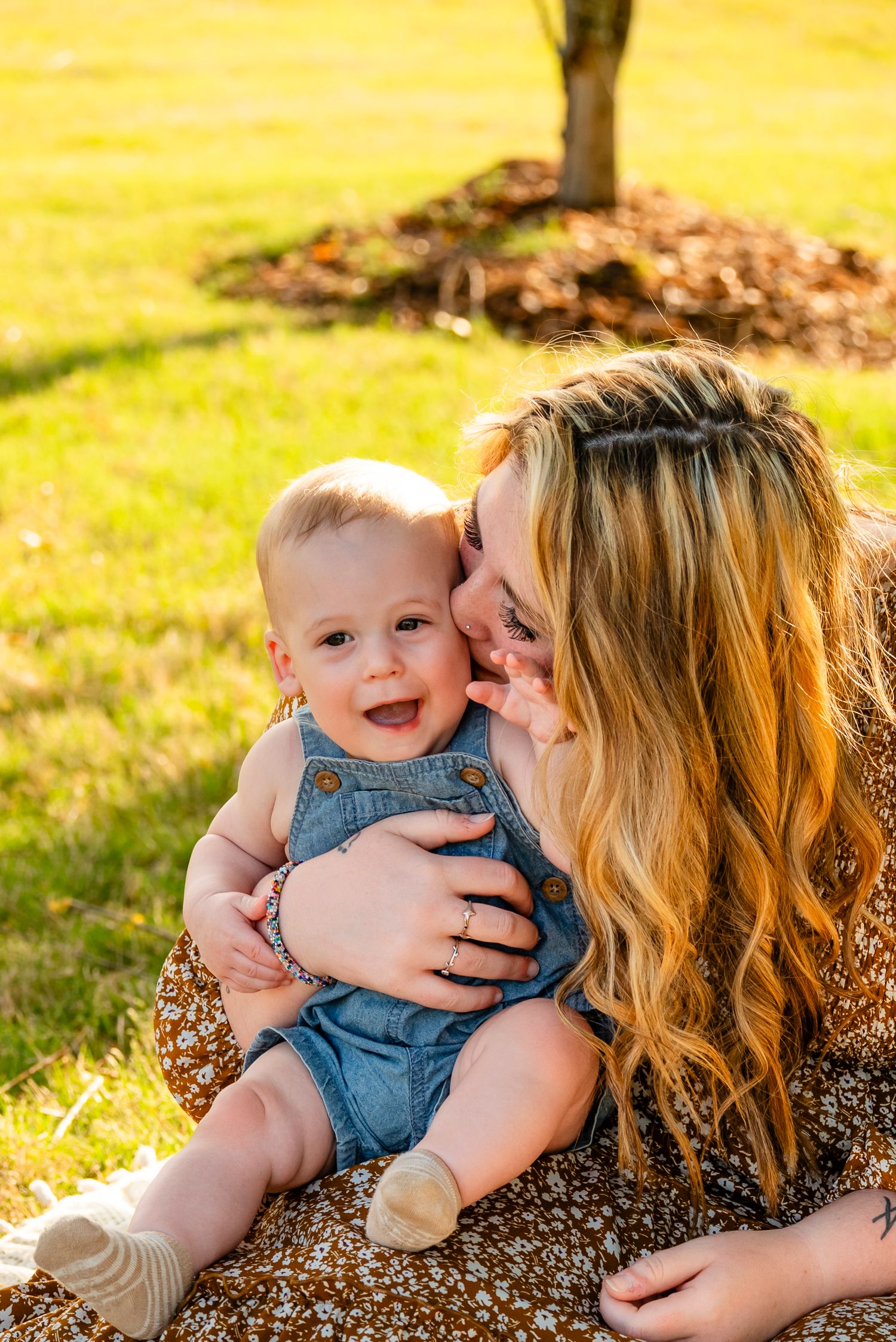 A smiling baby in denim overalls sits on a mother's lap outdoors on a sunny day.