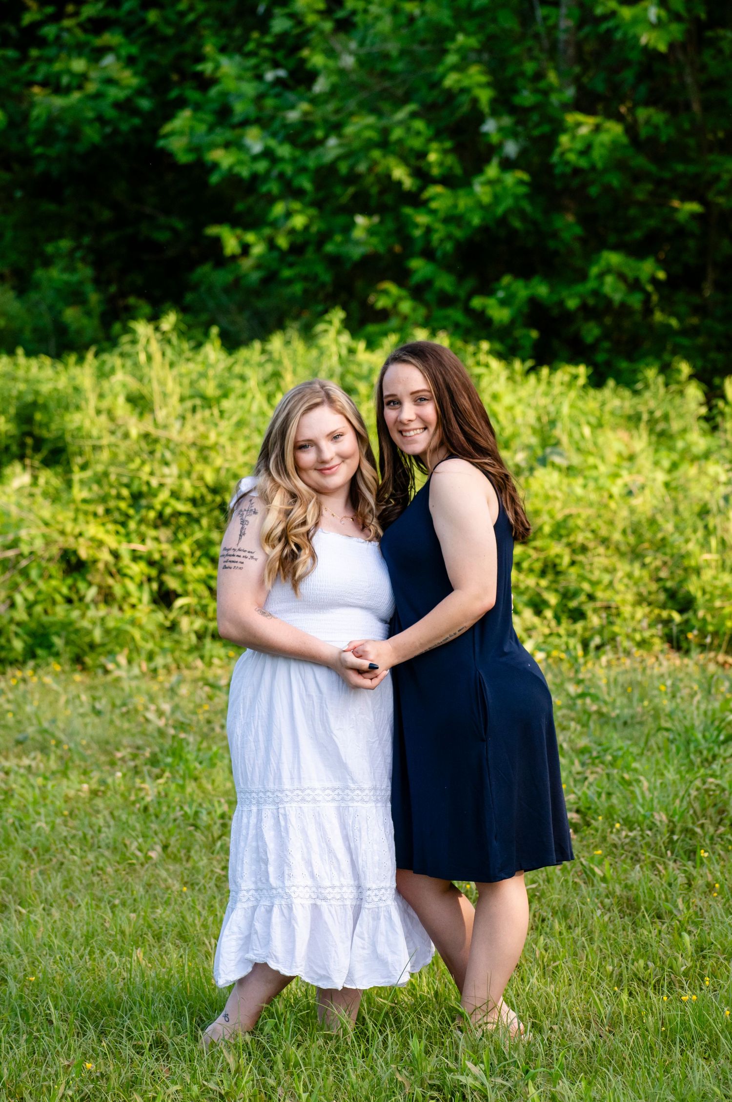 Two young women smile and embrace while standing together in a lush green outdoor setting.
