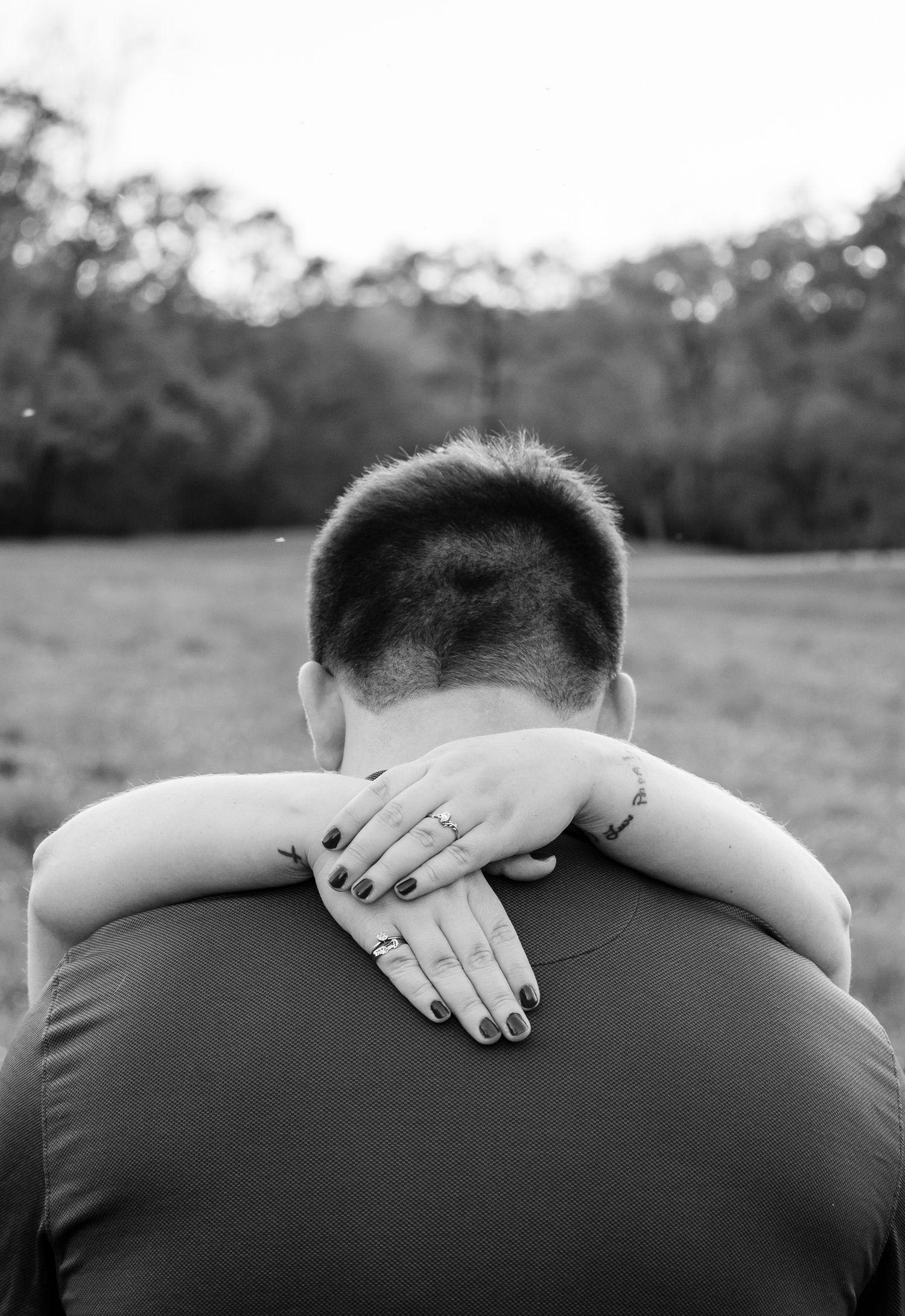 Black and white photo of a couple embracing outdoors, woman's hand with rings visible on man's neck.