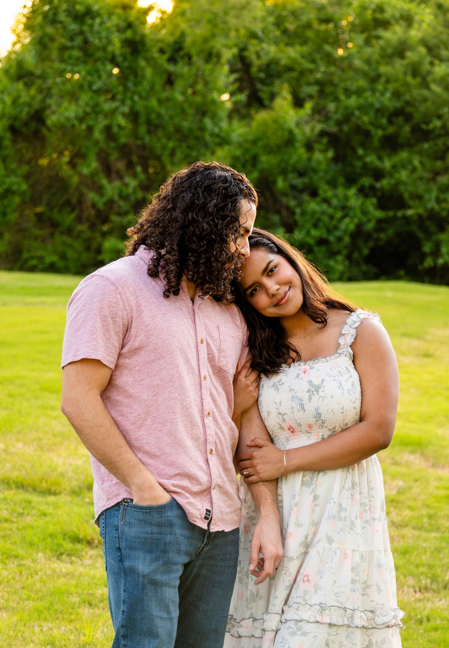 A couple embraces in a green park at golden hour, woman in floral dress leaning on man in pink shirt.