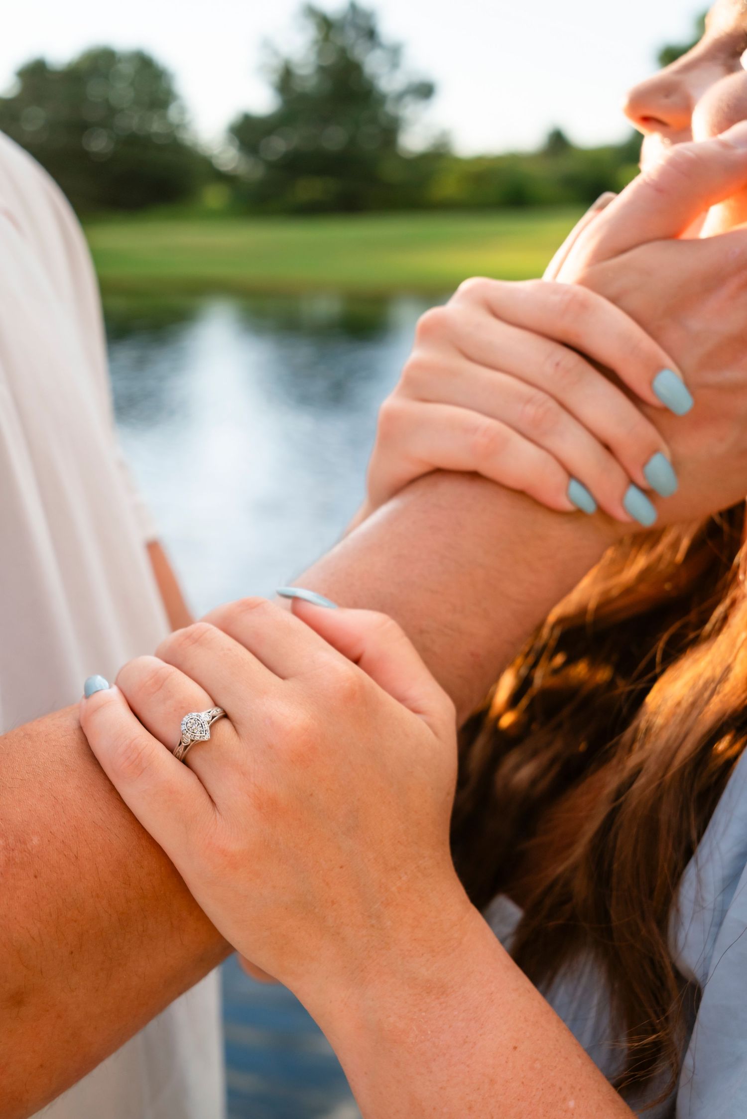 Close-up of a woman's hand wearing a diamond engagement ring while holding a man's arm near a lake.