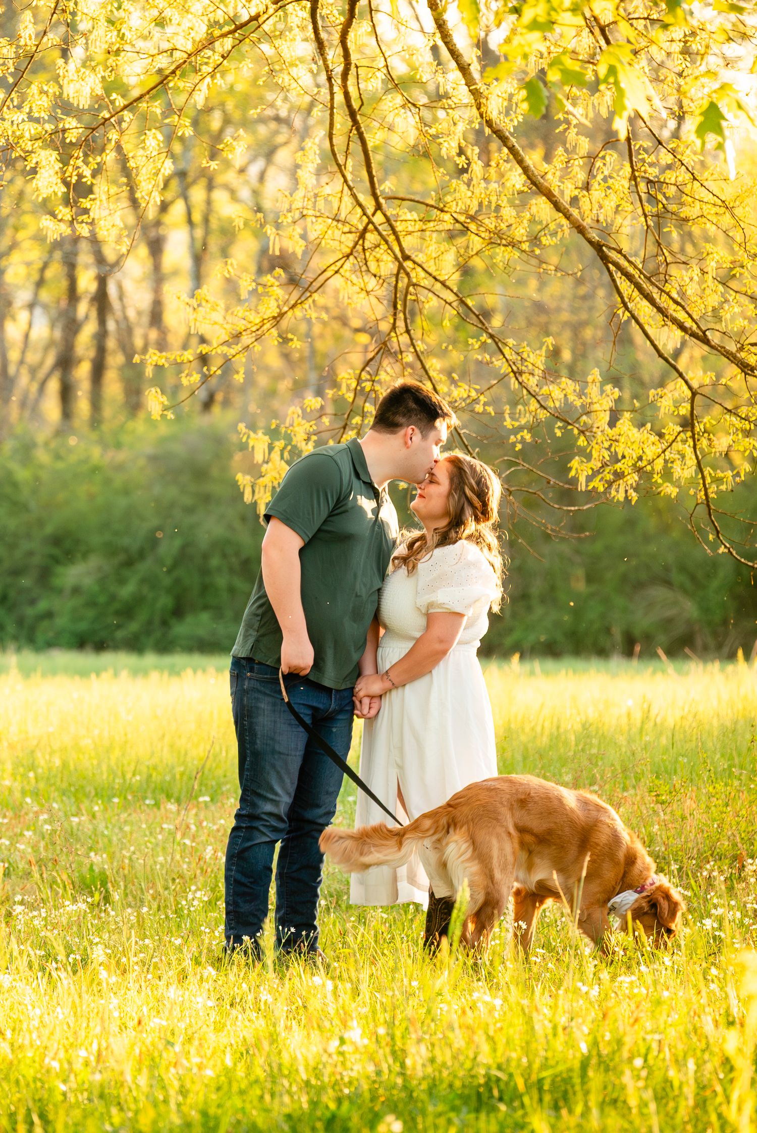 Couple sharing a romantic moment about to kiss in golden hour meadow with their dog on a leash.
