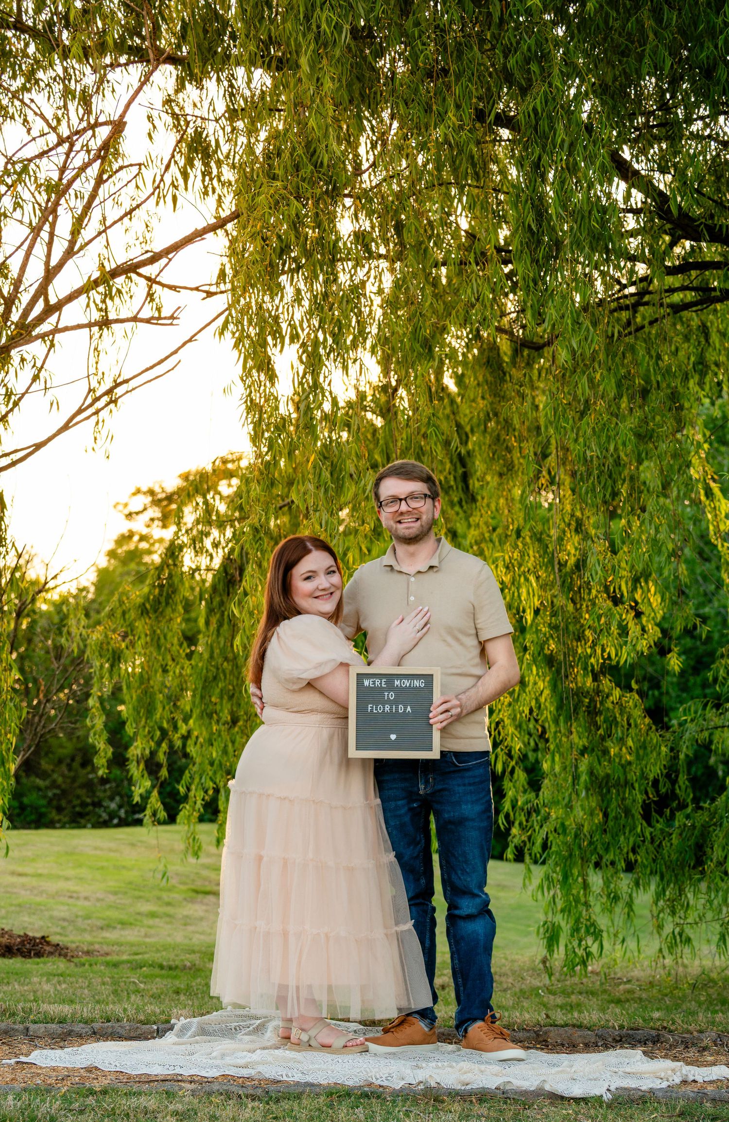 Smiling couple holding pregnancy announcement letterboard sign under weeping willow tree at golden hour outdoors.
