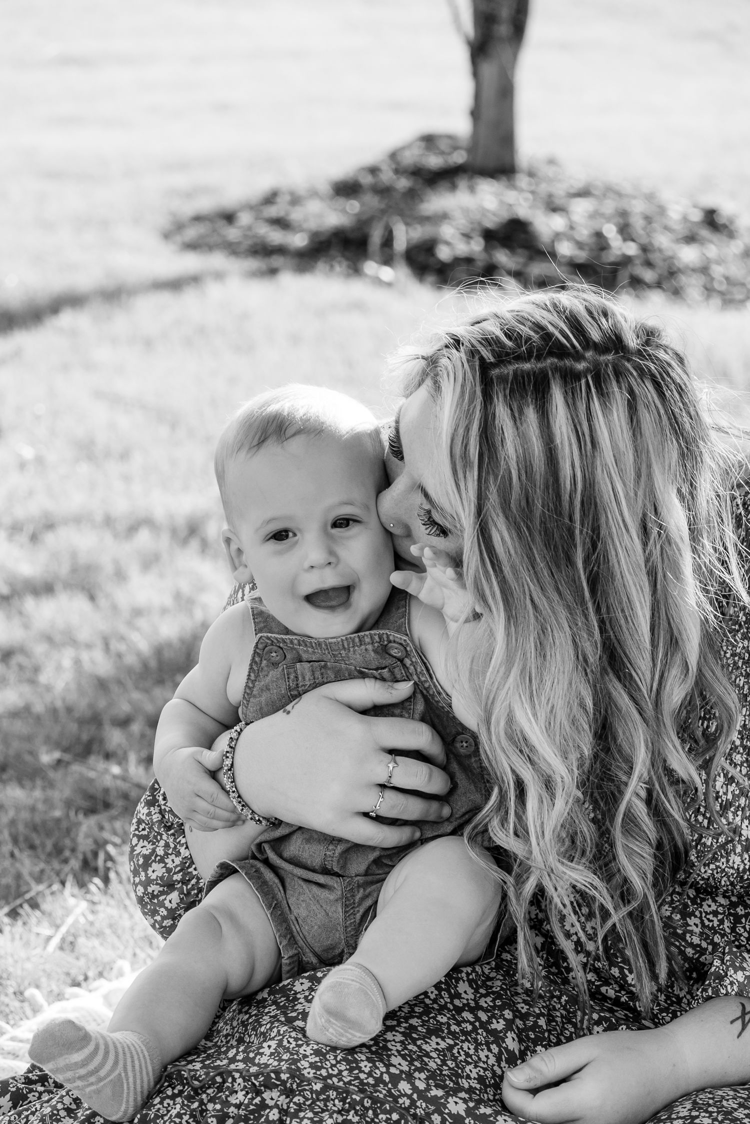 Black and white photo of a smiling baby in overalls sitting on a woman's lap outdoors.