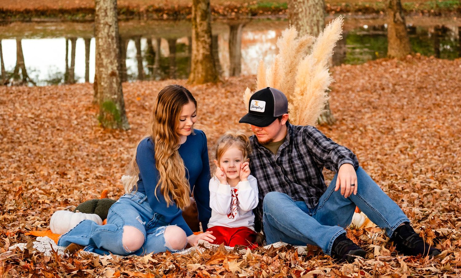 Young family with toddler girl sitting together in colorful autumn leaves near a pond.