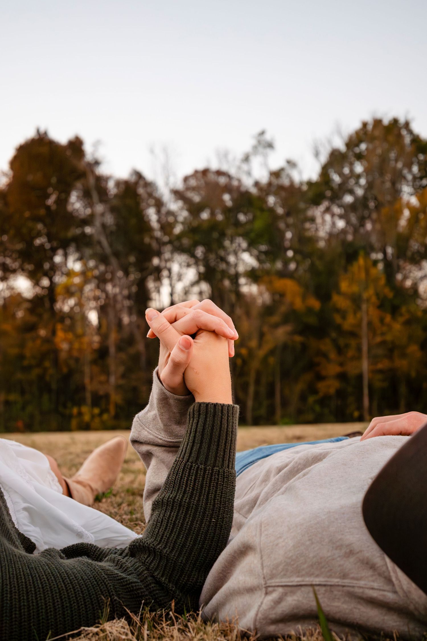 Close-up of couple holding hands while lying on grass with autumn trees and sky in background.