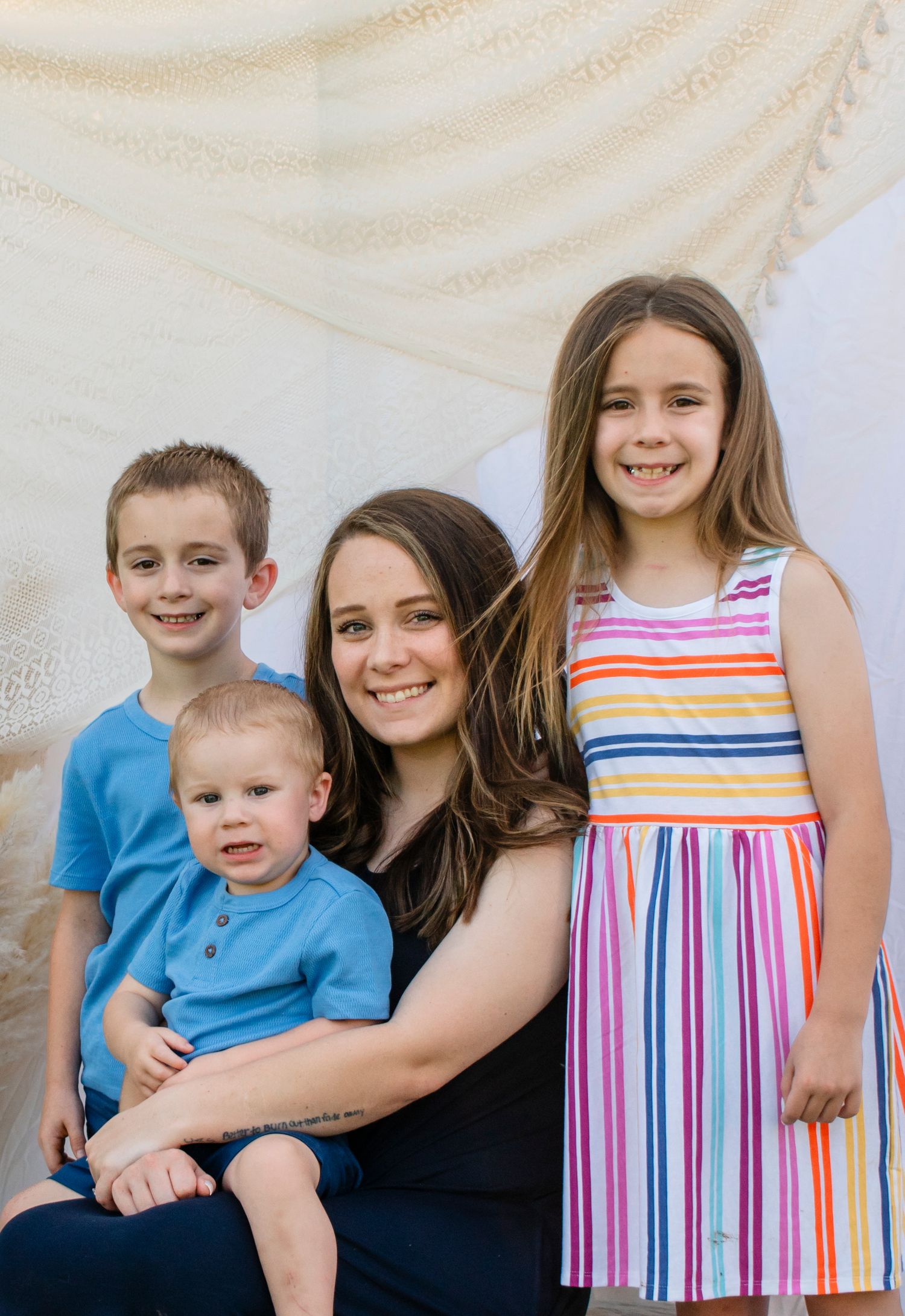 A smiling mother sits with her three children, two boys in blue shirts and a girl in a colorful striped dress.