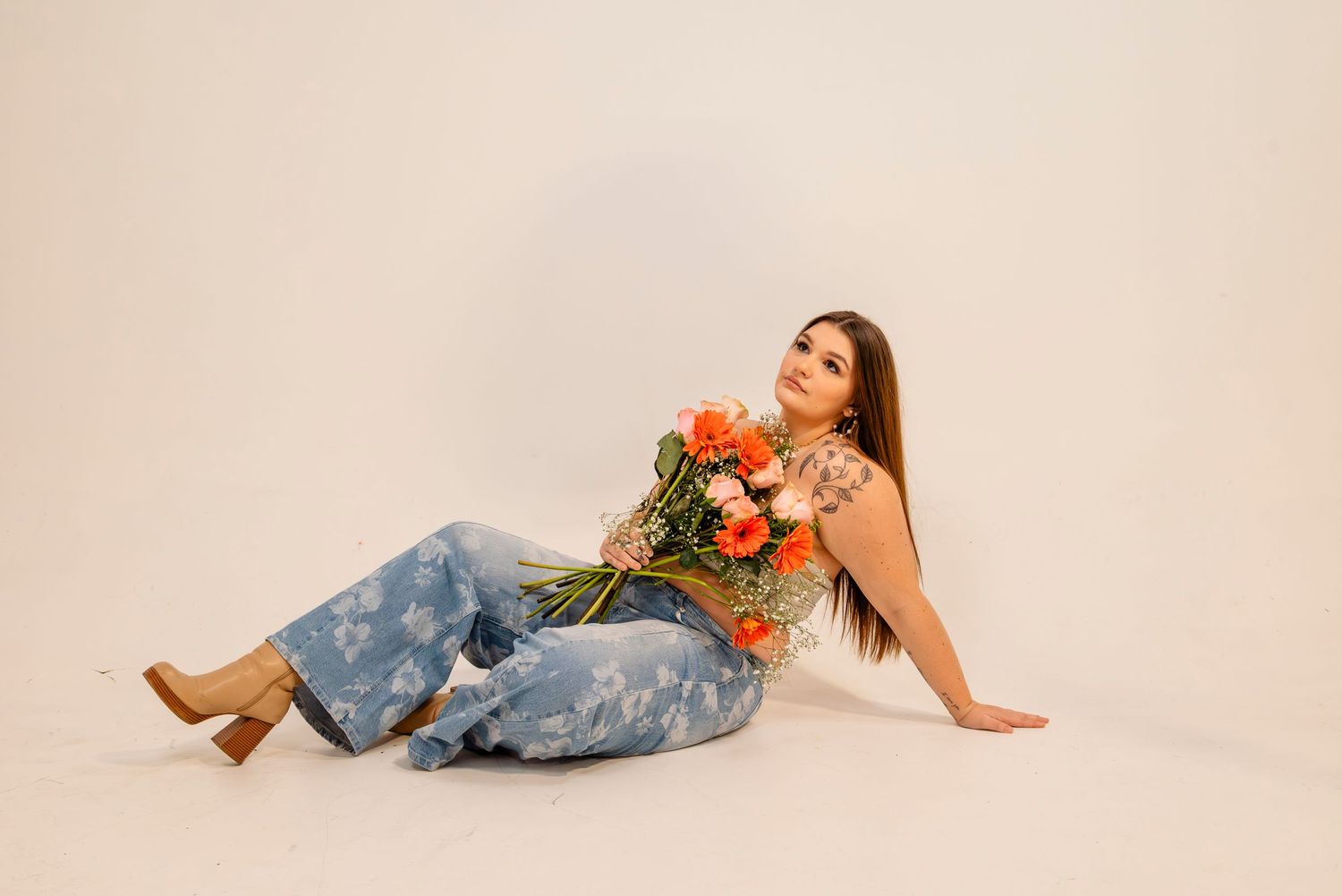 Woman with tattoos sits on floor in jeans holding a vibrant orange and pink floral bouquet.