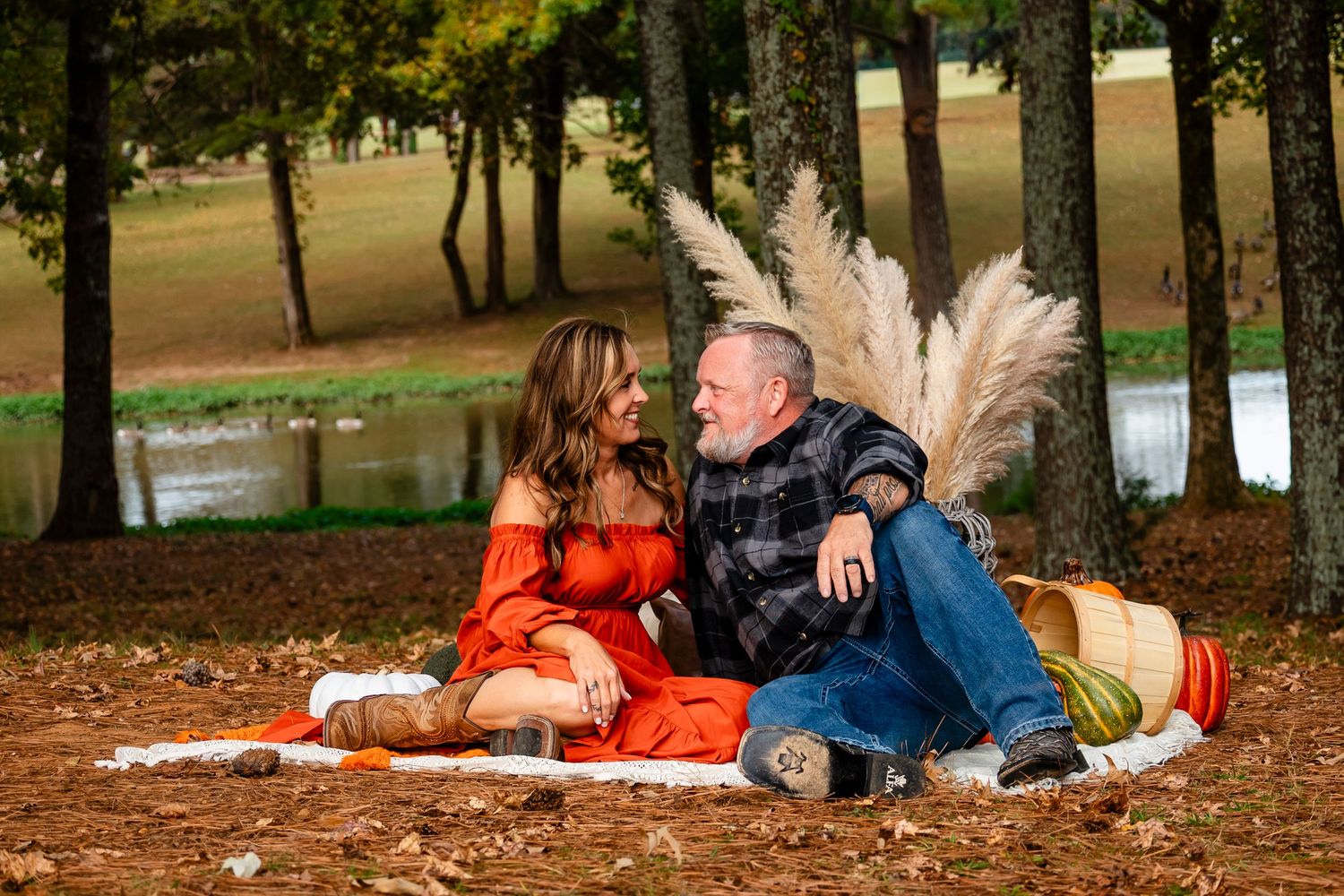 Couple sitting together on fall leaves near pond with pampas grass decor.