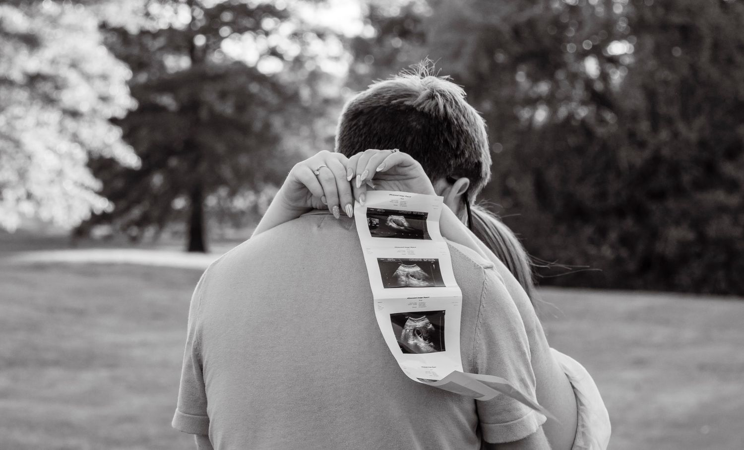 Black and white photo of a couple embracing outdoors, holding ultrasound photos, announcing their pregnancy.