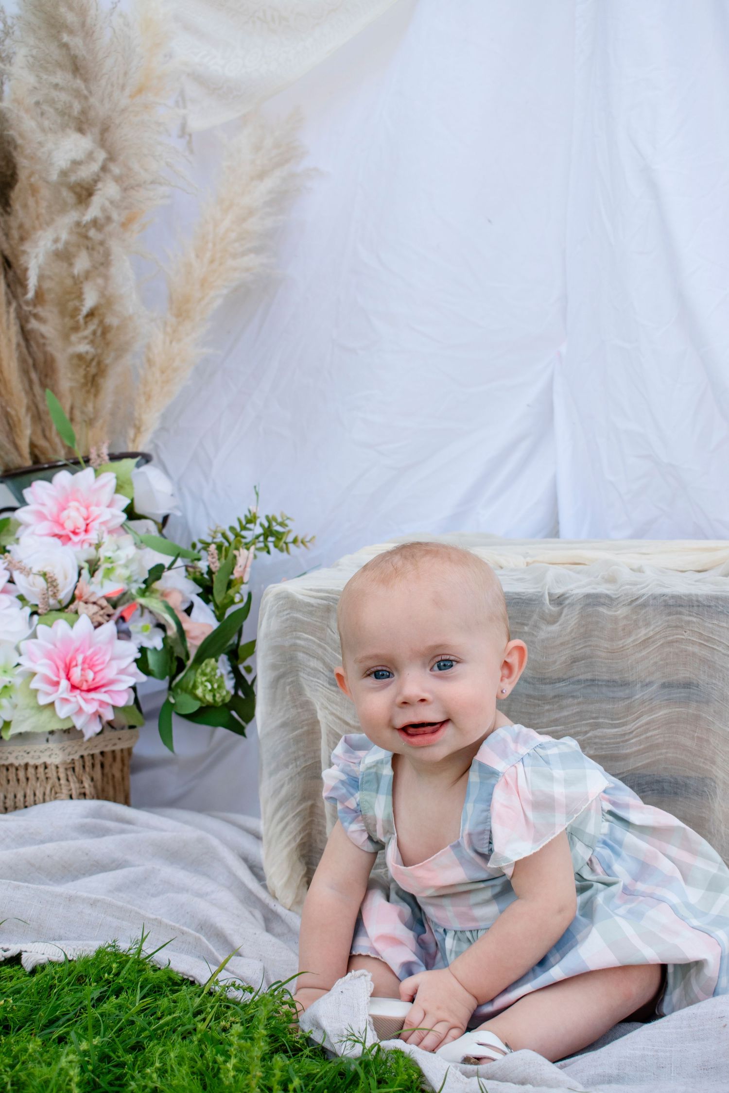Adorable baby girl in pastel dress sitting near pink flowers and pampas grass in a bright studio setting.
